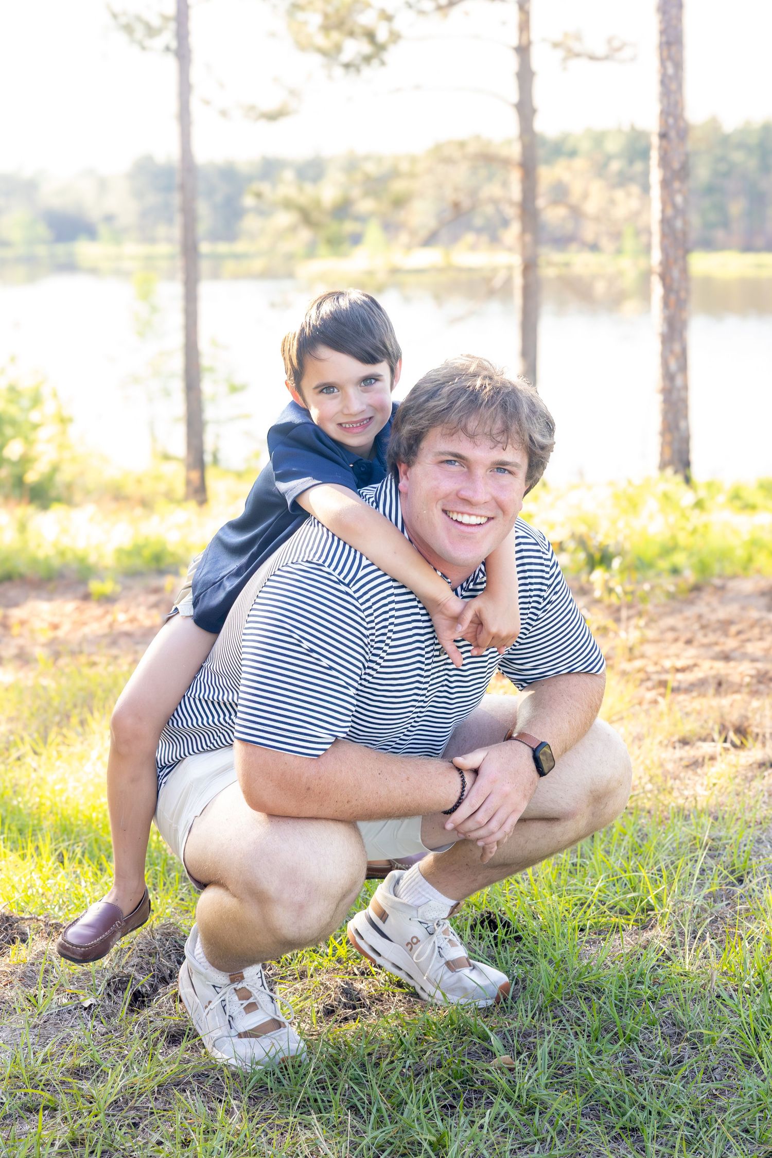A smiling young man crouches outdoors as a boy rides on his shoulders near a sunlit lake surrounded by pine trees.