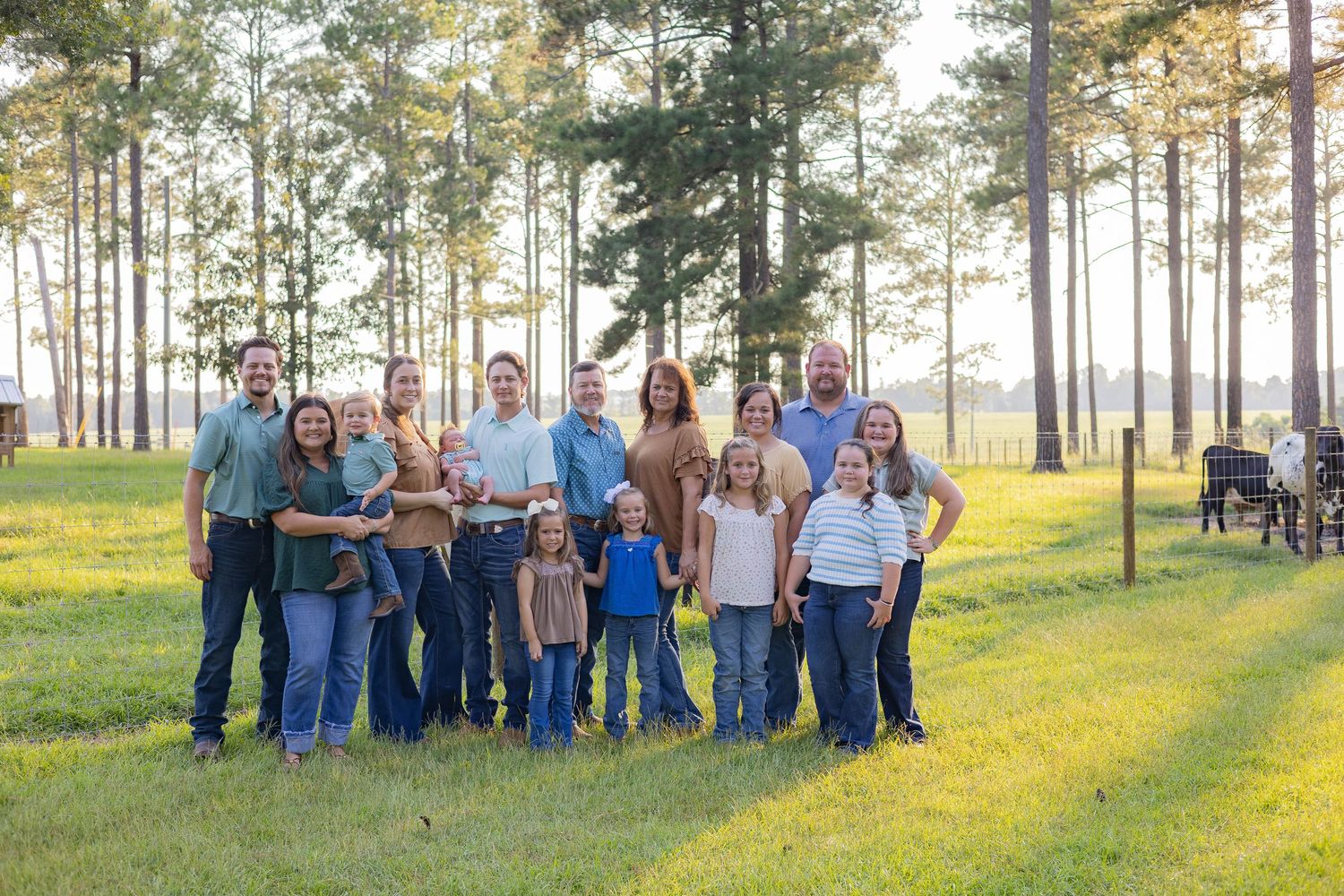Large family group portrait outdoors in a sunlit pine forest clearing, posing together on green grass.