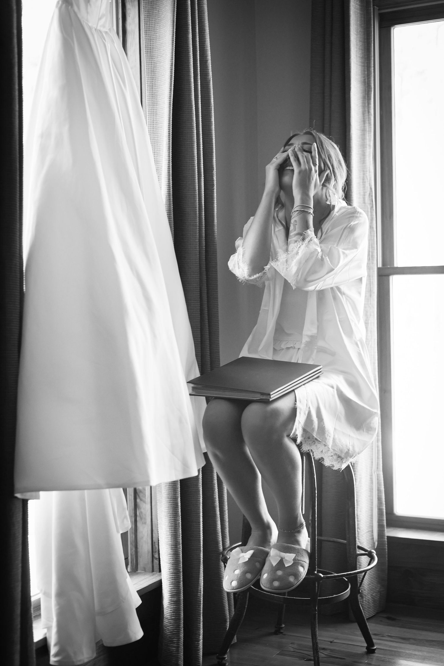 Black and white photo of a bride in white robe sitting on stool near window with wedding dress hanging nearby.