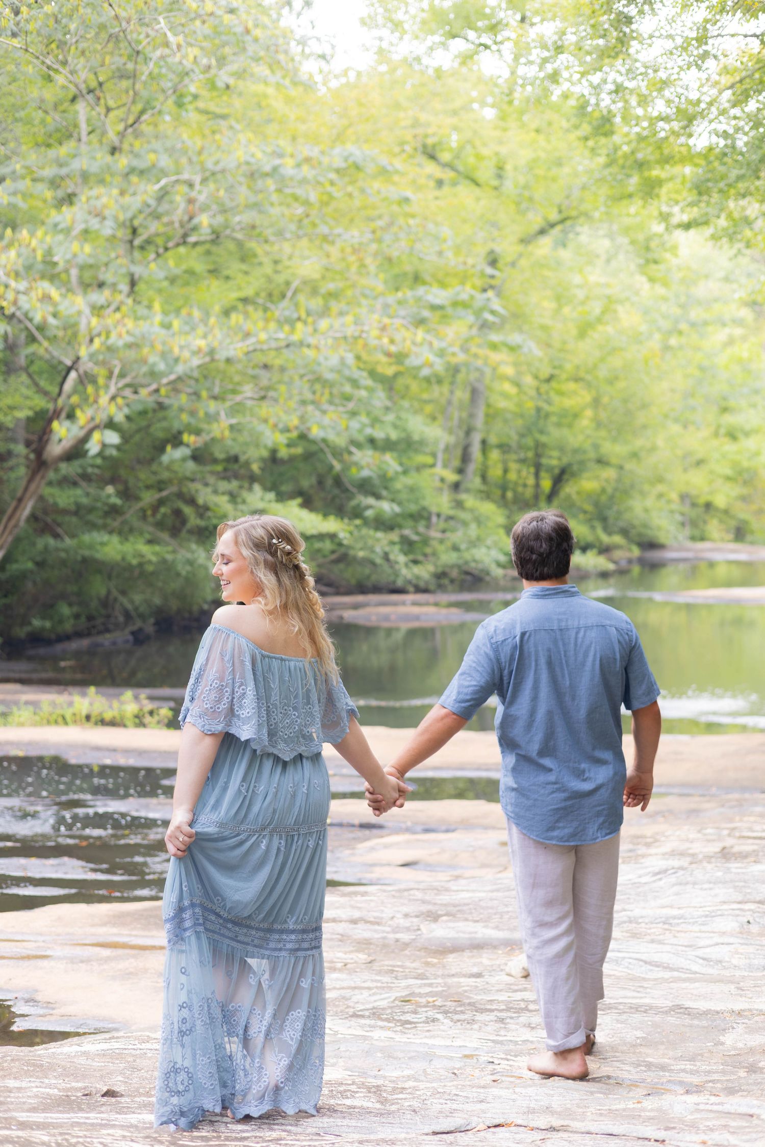 A couple holding hands walks toward a serene river surrounded by lush green trees, woman in a flowing blue lace dress.
