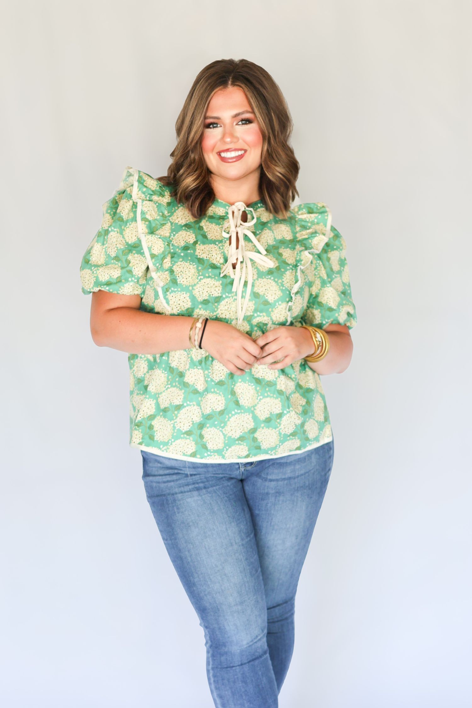 Woman smiling in green floral puff sleeve top with ruffle tie neck detail and blue jeans.