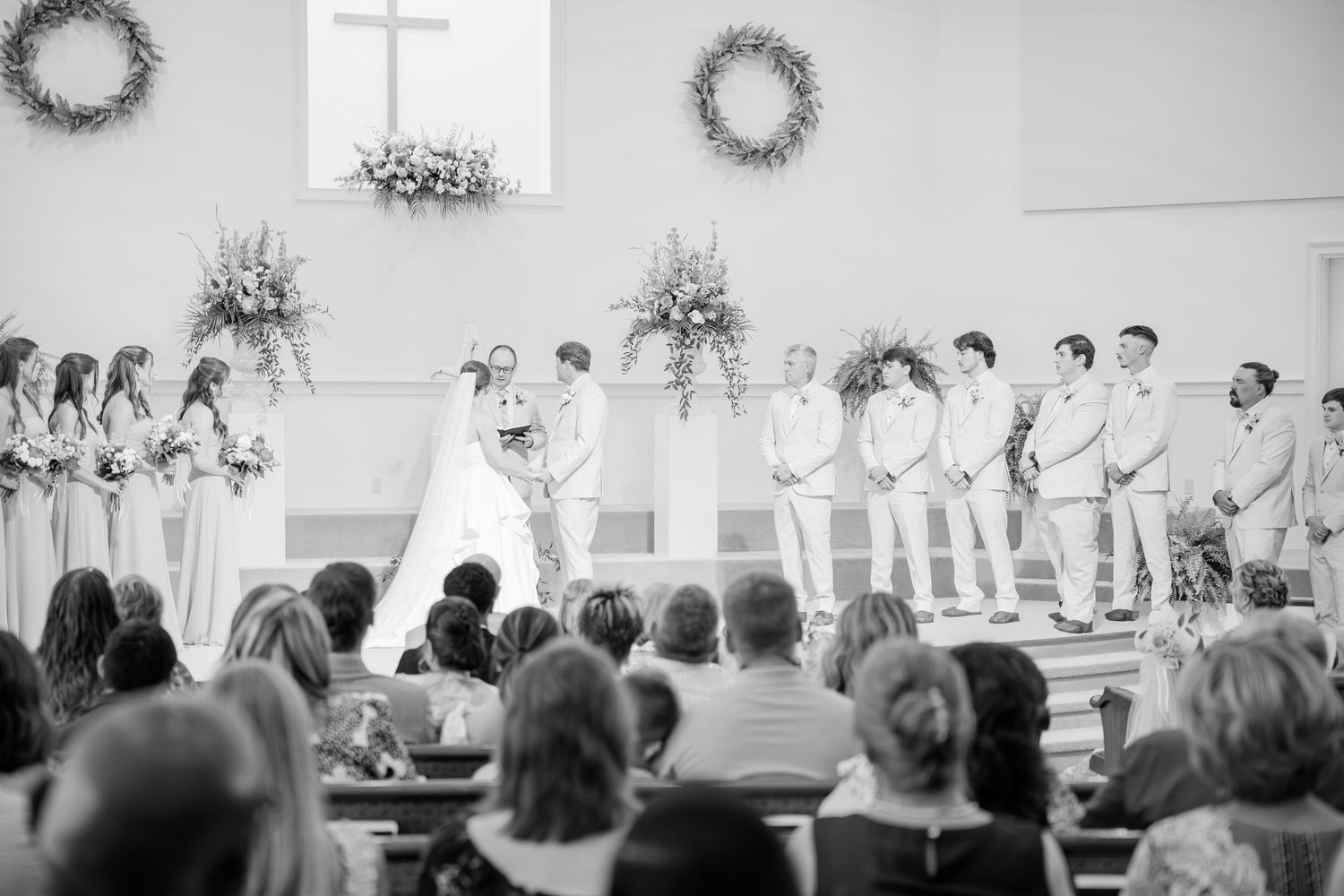 Black and white wide shot of wedding ceremony with bridal party at the altar of a decorated church.
