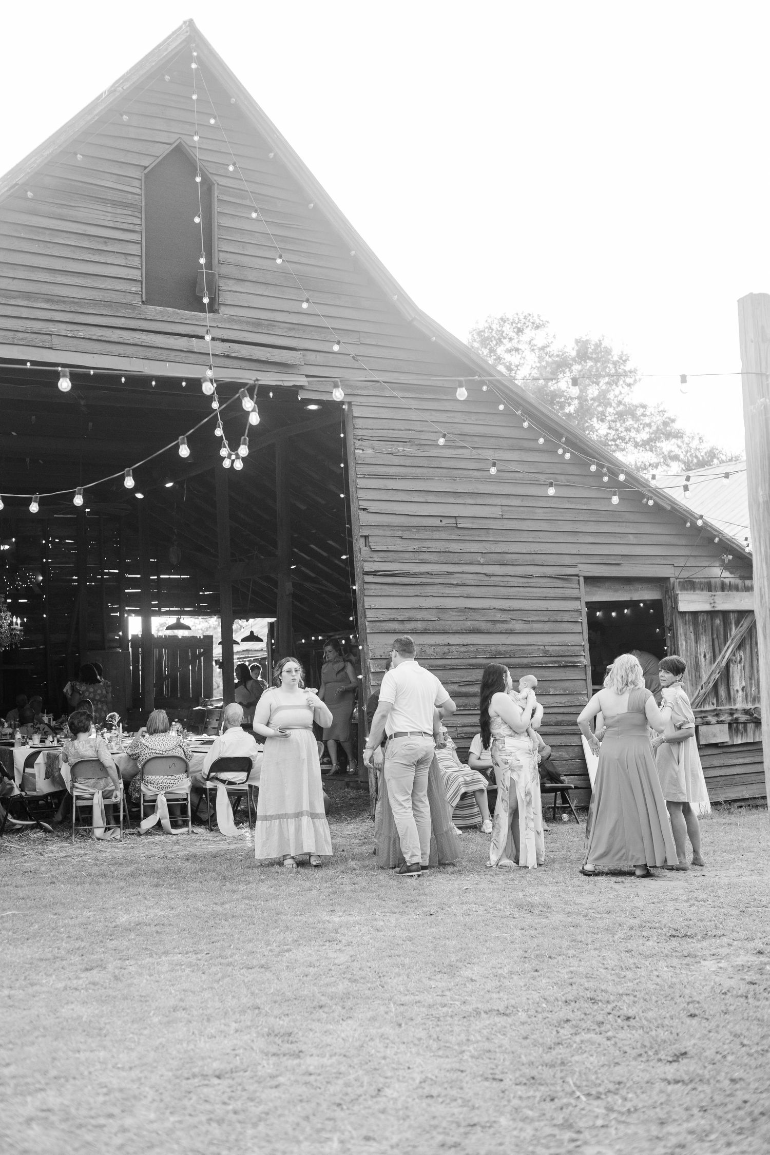 Black and white photo of wedding guests mingling outside a rustic barn venue decorated with string lights.