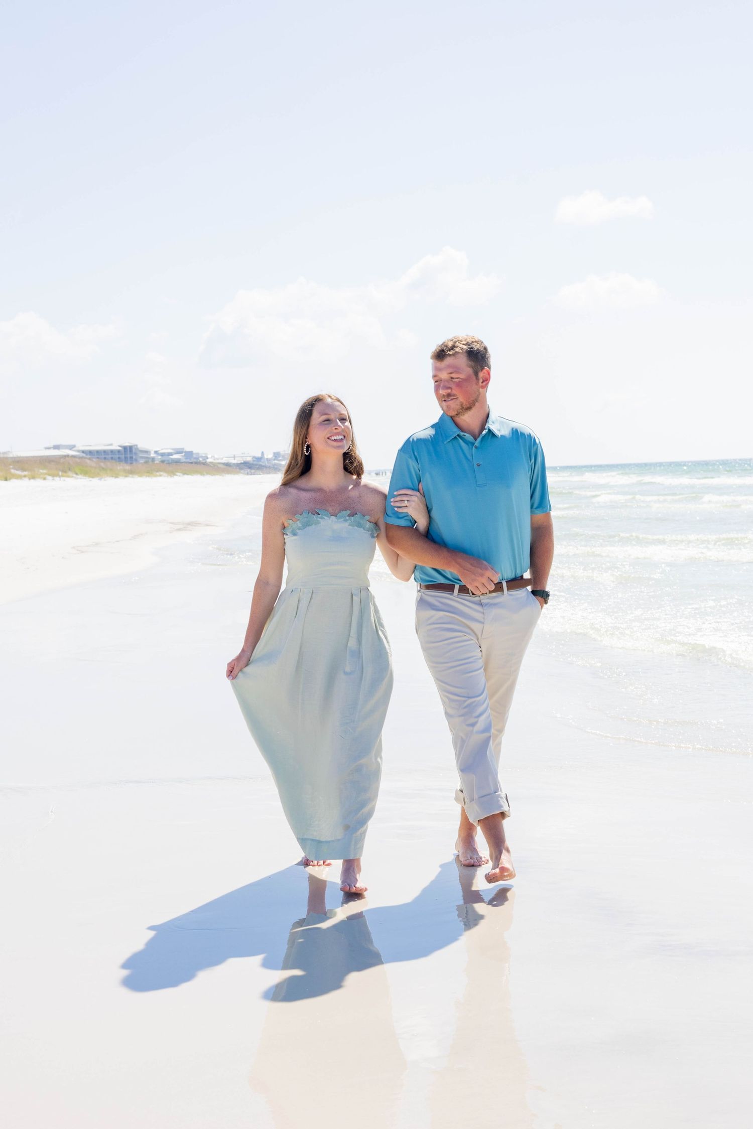 Couple walking hand in hand along a bright sandy beach with ocean waves and blue sky.