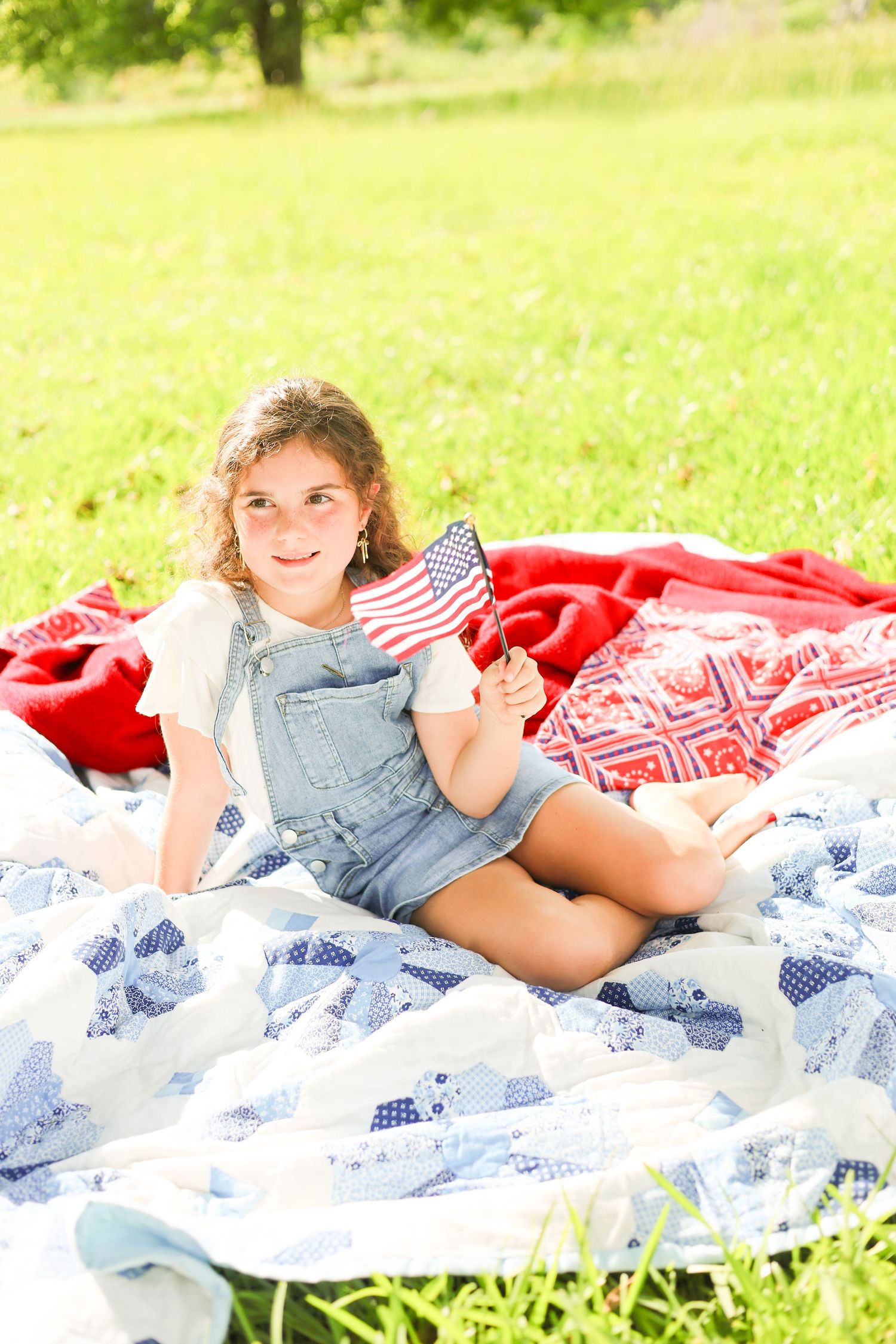 Young girl sitting on a quilt in a park, holding a small American flag, wearing denim overalls on a sunny day.