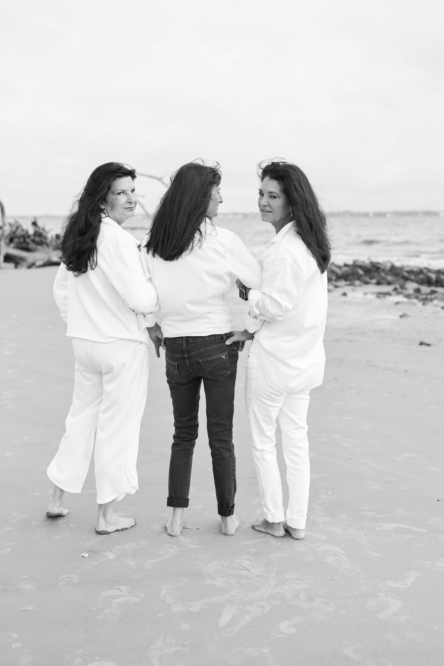 Three friends in white outfits and dark hair standing together on a beach in black and white photography.