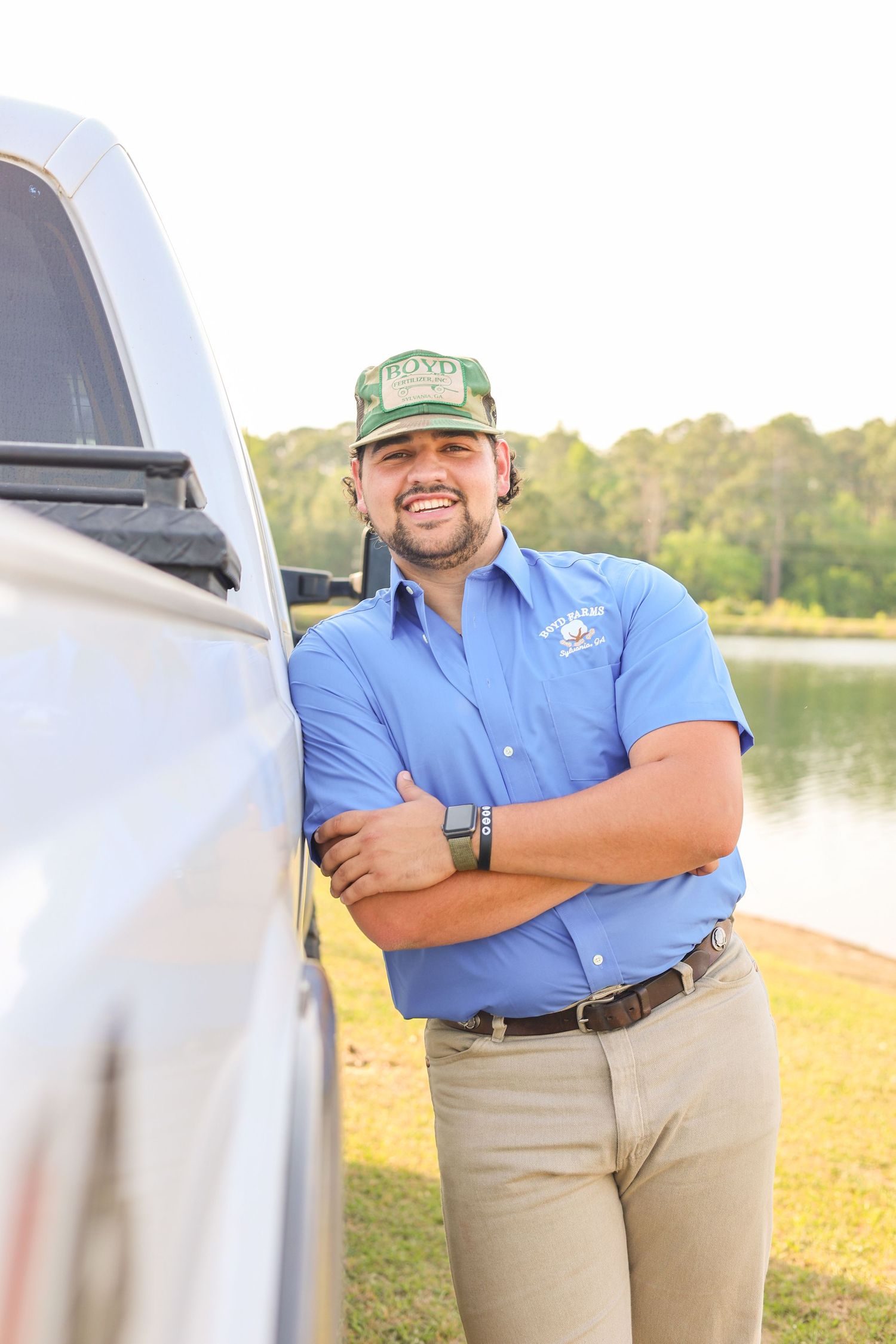 Person in work uniform and green cap stands next to white truck by lakeside.