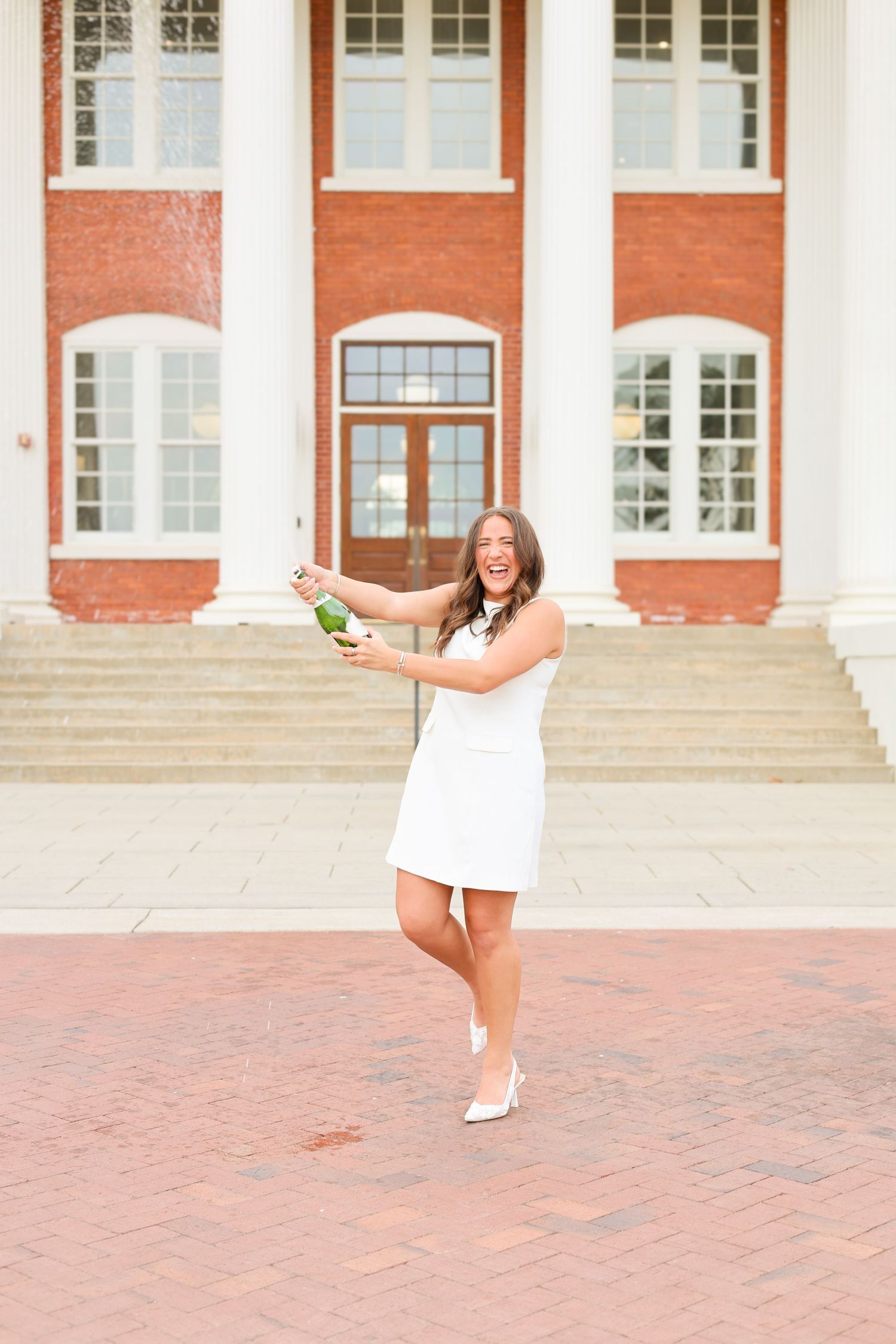 Person in white dress celebrating with champagne on brick courtyard in front of red brick building.