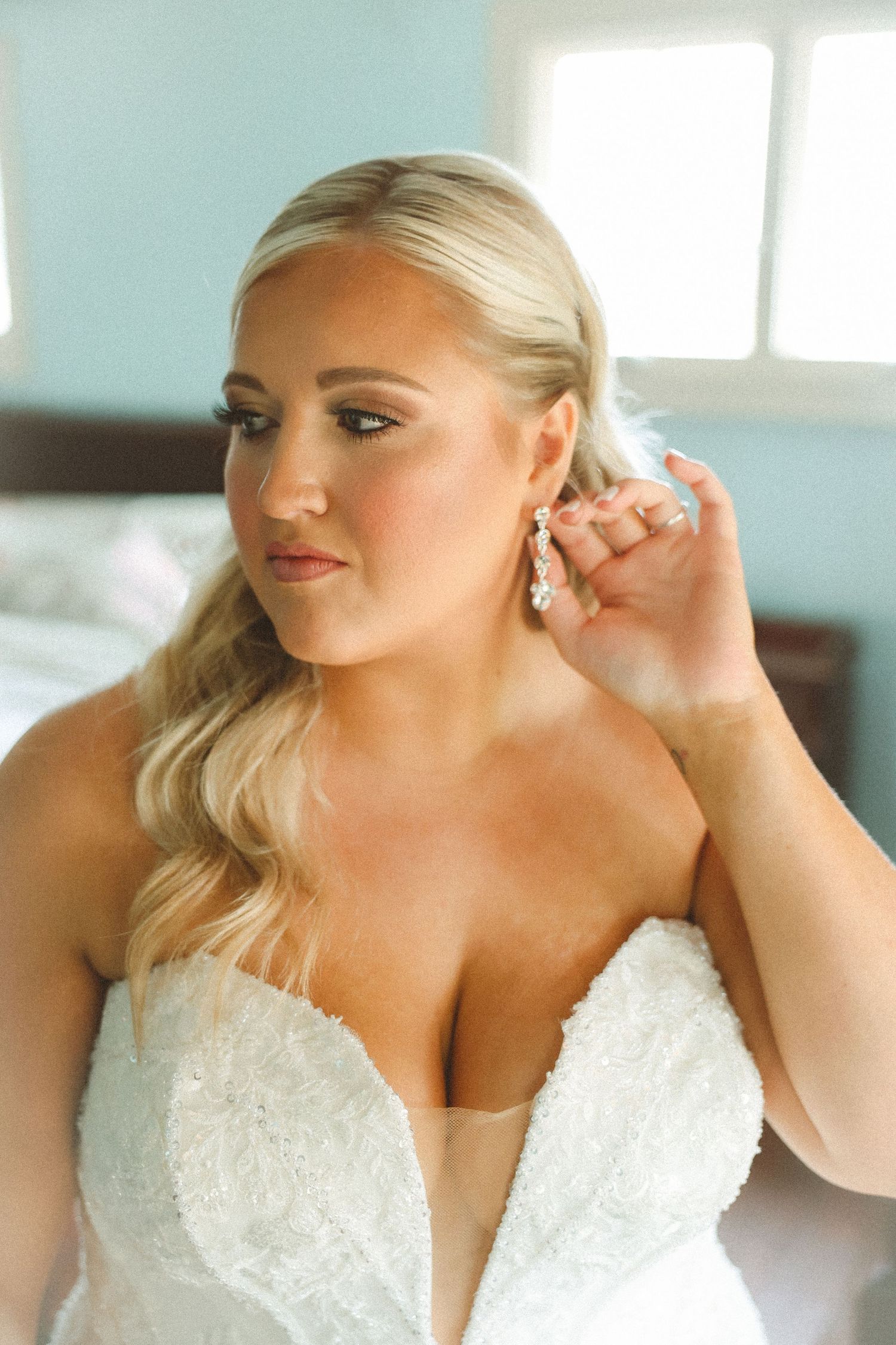 Bride in strapless lace wedding gown adjusting elegant drop earring, with soft glam makeup and blonde waves.