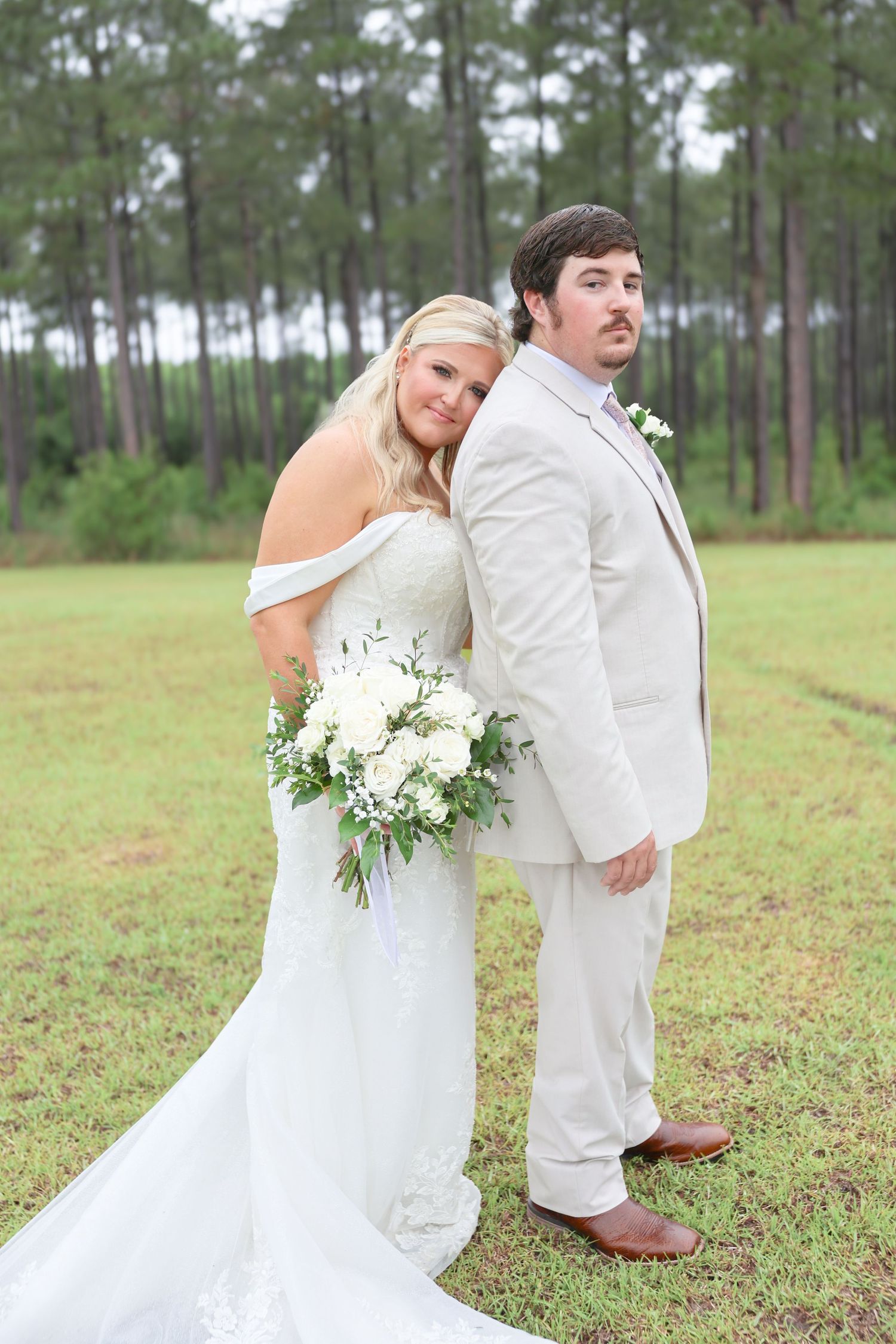 Bride and groom pose back-to-back in a field, bride in off-shoulder lace gown holding white floral bouquet.