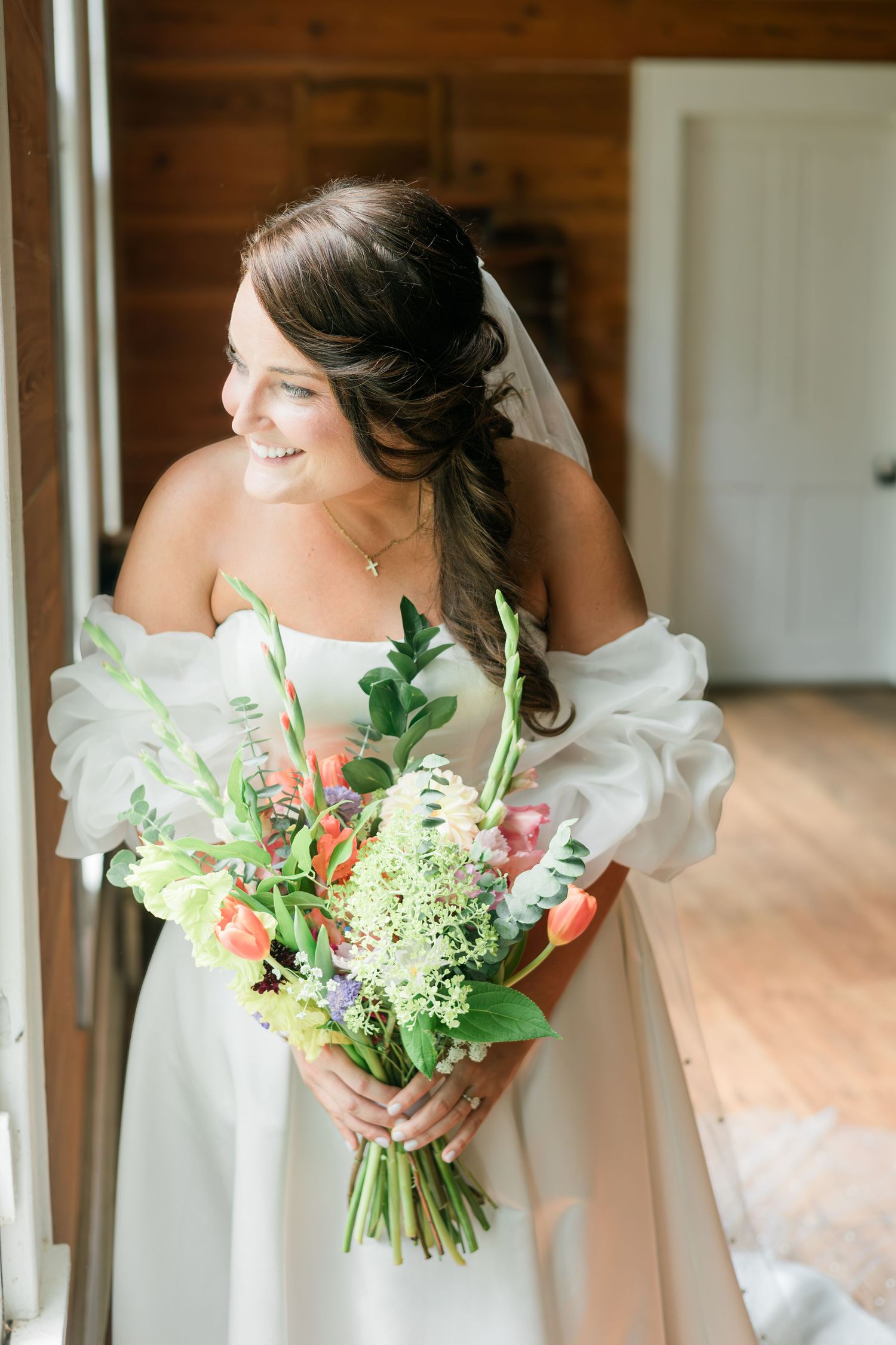 Smiling bride in white off-shoulder gown holding colorful spring bouquet with tulips and greenery in rustic wood interior.