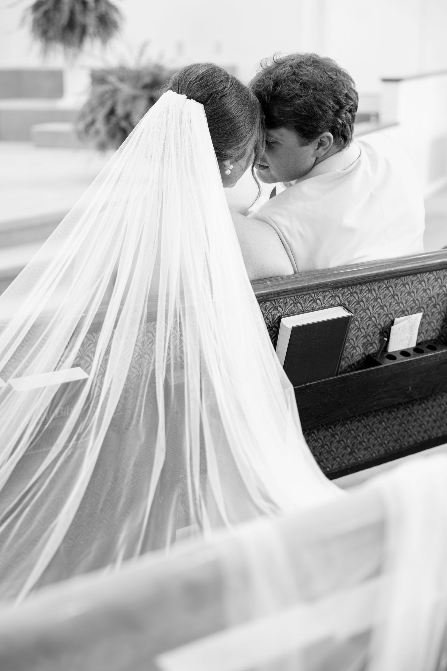 Bride and groom sharing intimate moment in church pew with flowing veil draped over wedding dress.