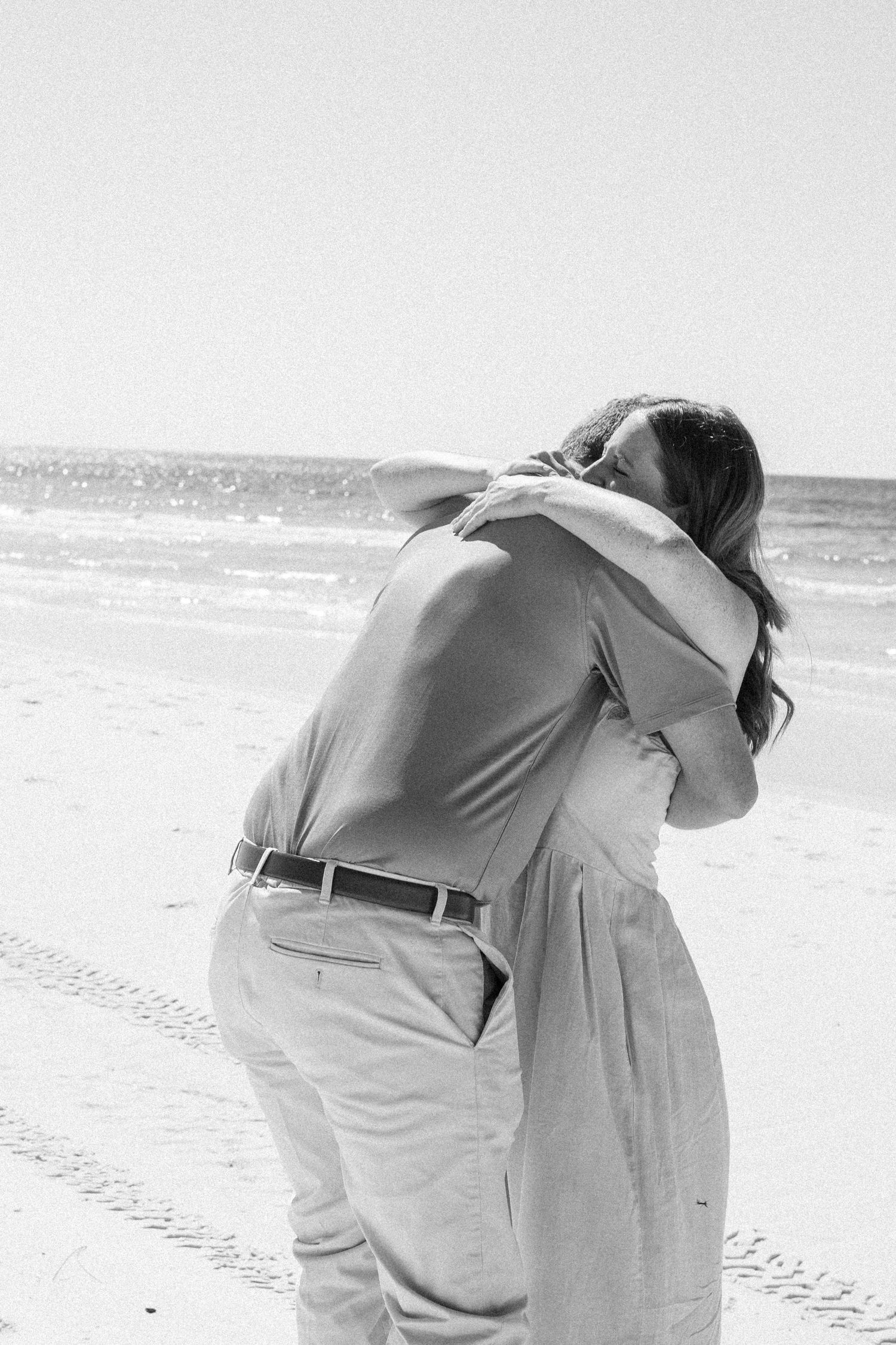 Black and white photo of a couple embracing on a sandy beach with ocean waves in the background.