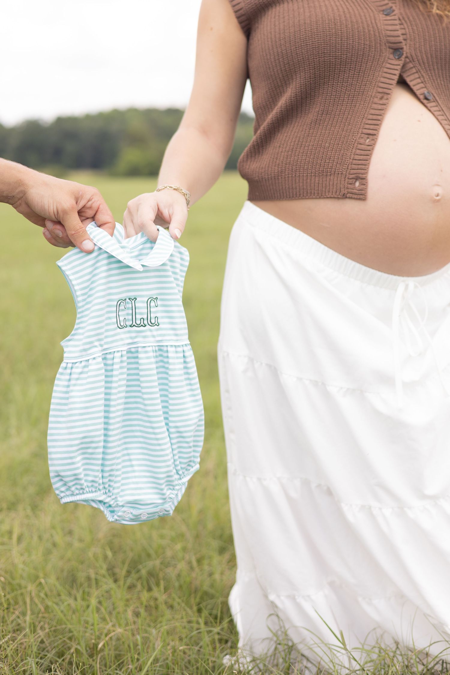 Pregnant woman in white skirt holding a monogrammed blue striped baby romper outdoors in a green field.