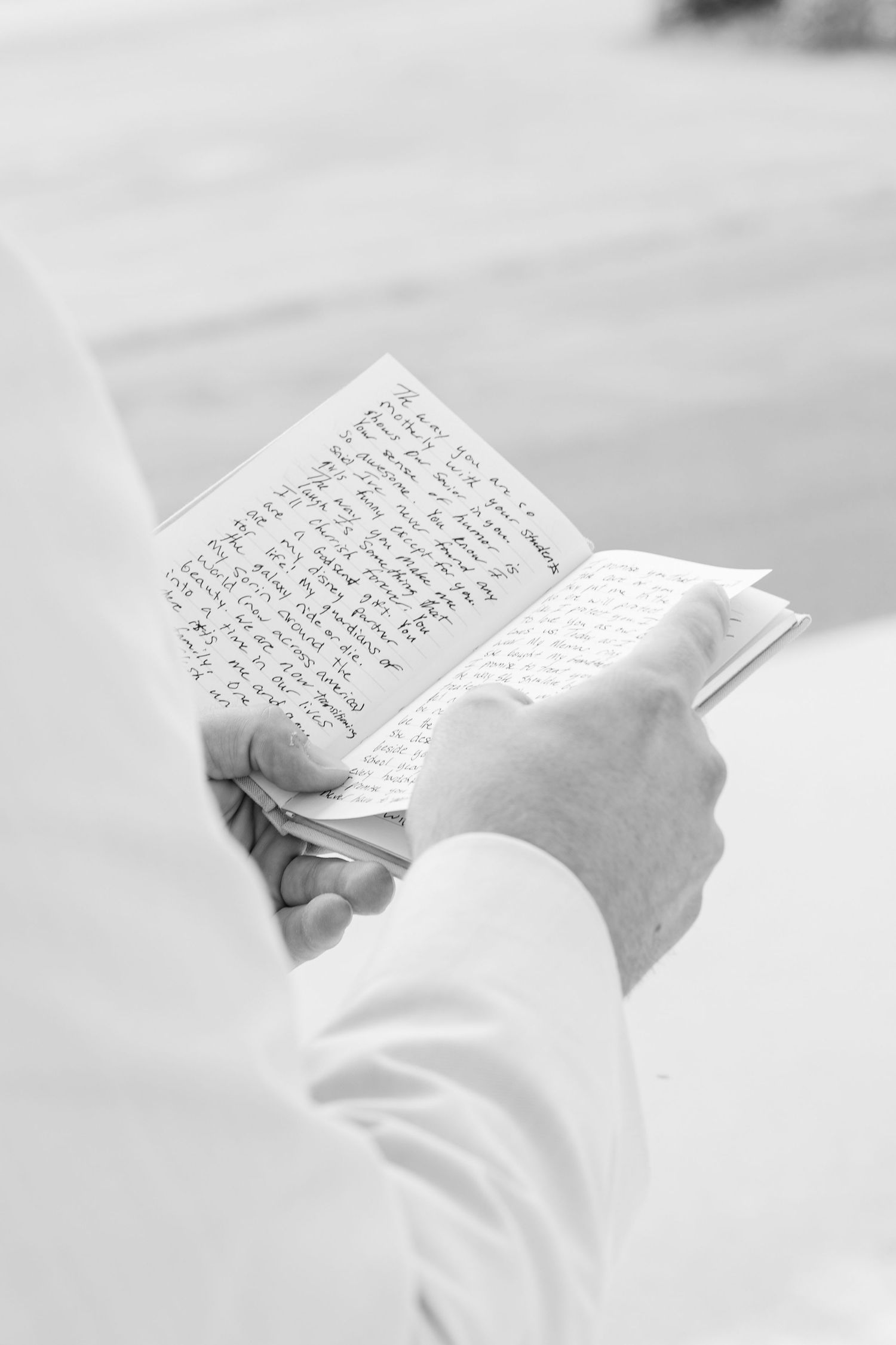 Person in white clothing reading a small handwritten book outdoors, black and white photography.