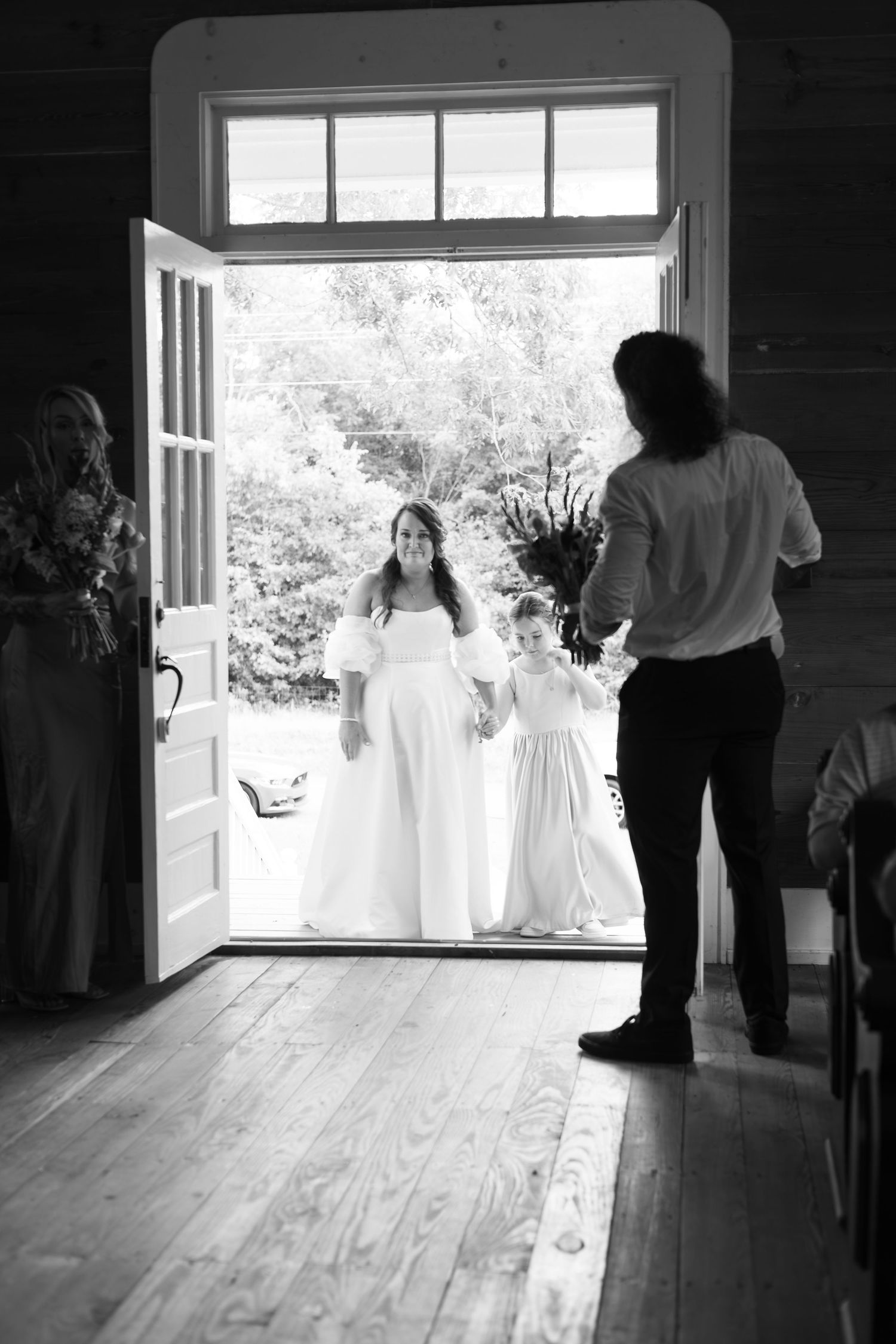 Black and white photo of a bride in white gown with flower girl entering church doors, greeted by a guest inside.