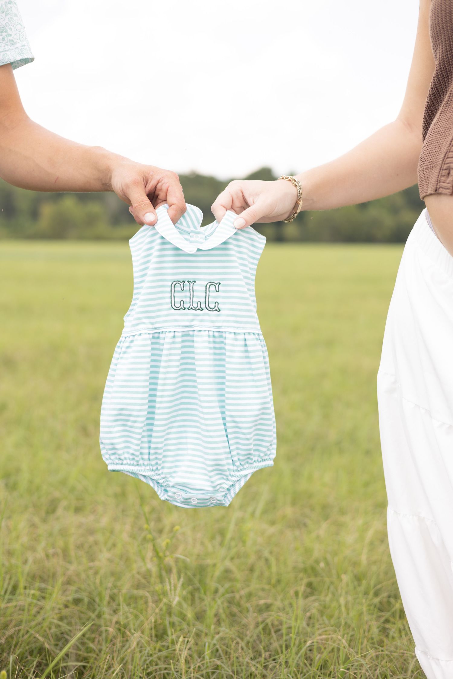 Two people holding a monogrammed blue striped baby romper outdoors in a green field.