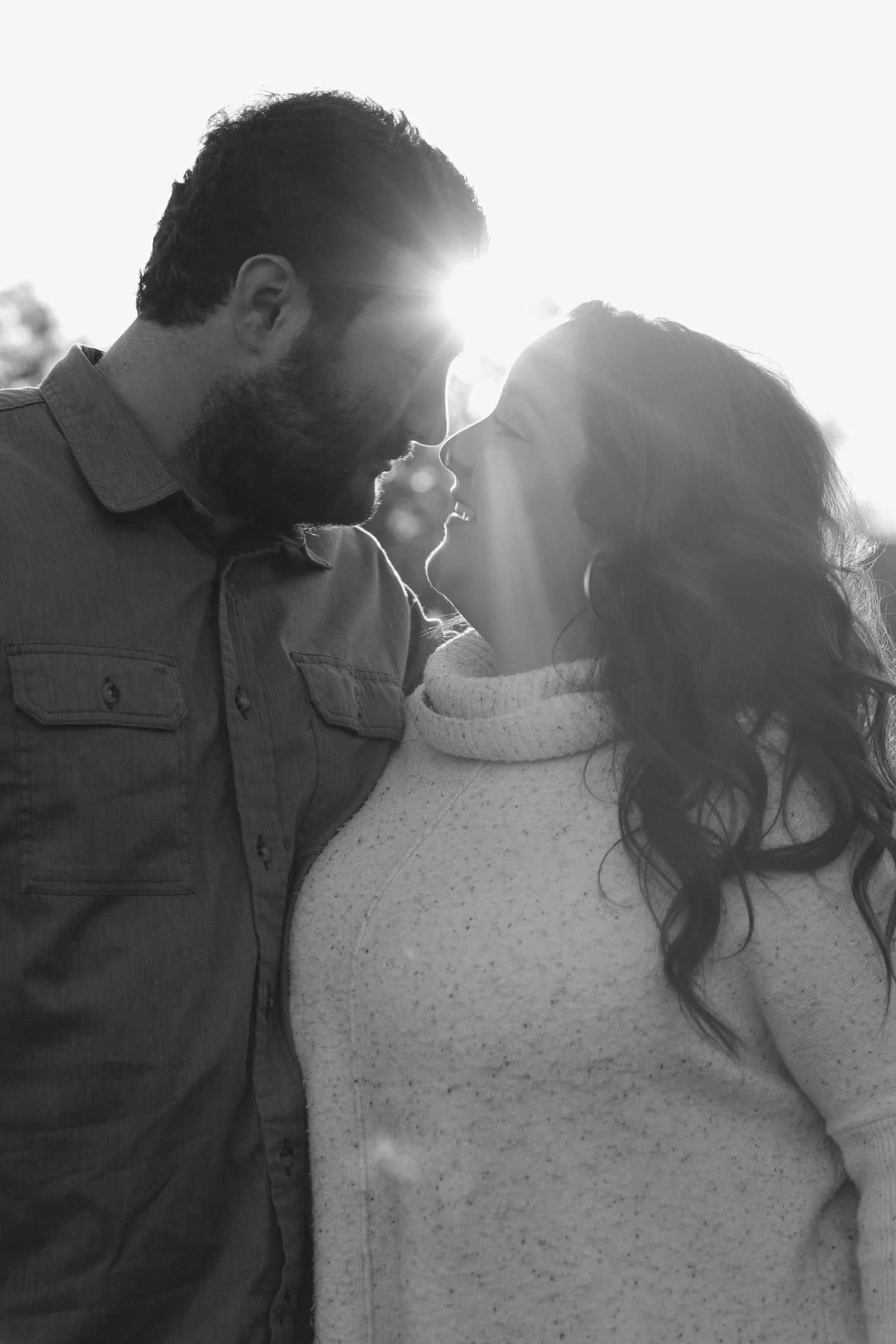 Black and white photo of a romantic couple almost kissing with sunlight glowing between them outdoors.
