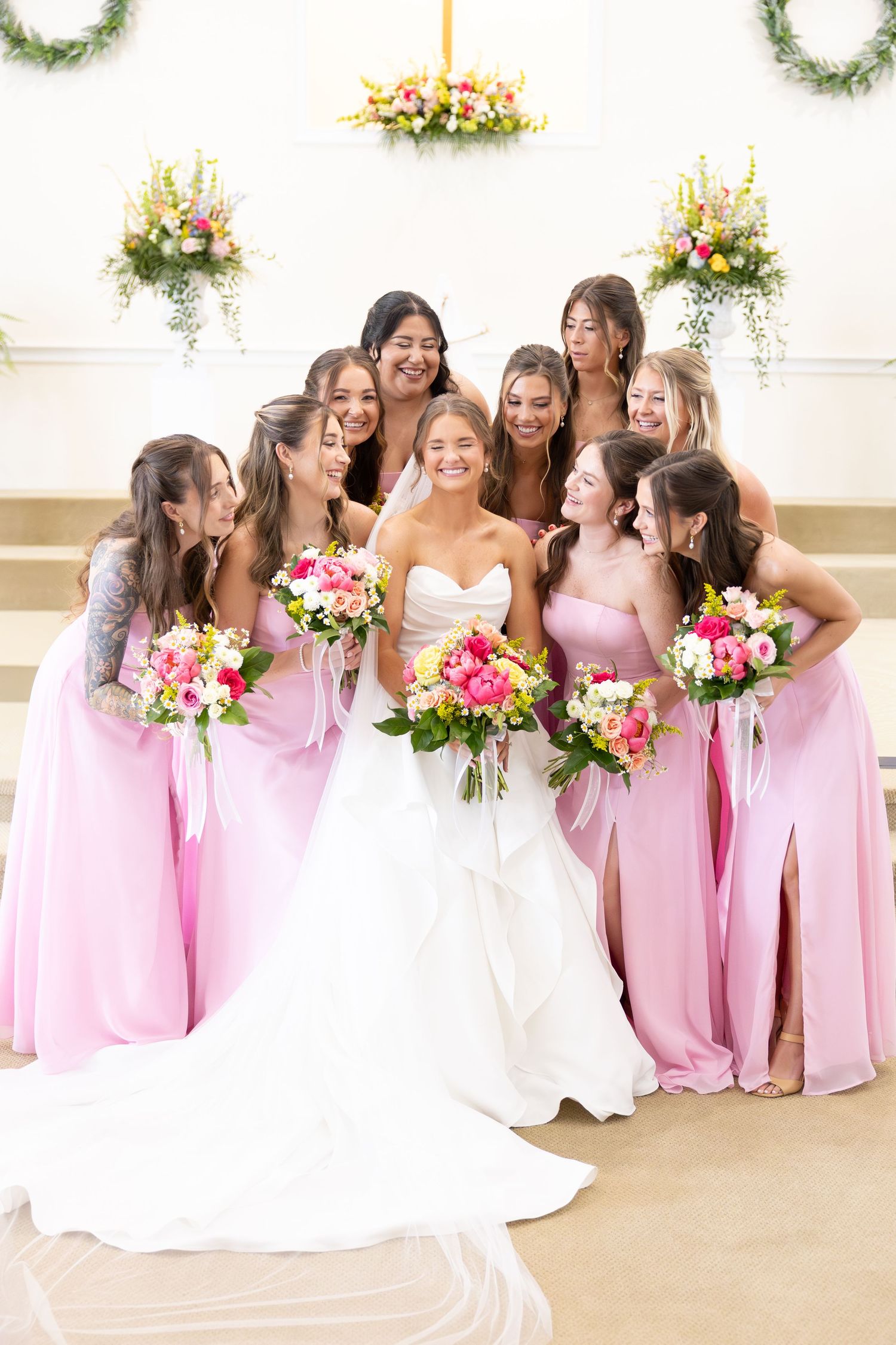 Bride in white gown surrounded by bridesmaids in pink dresses holding colorful bouquets inside a decorated chapel.