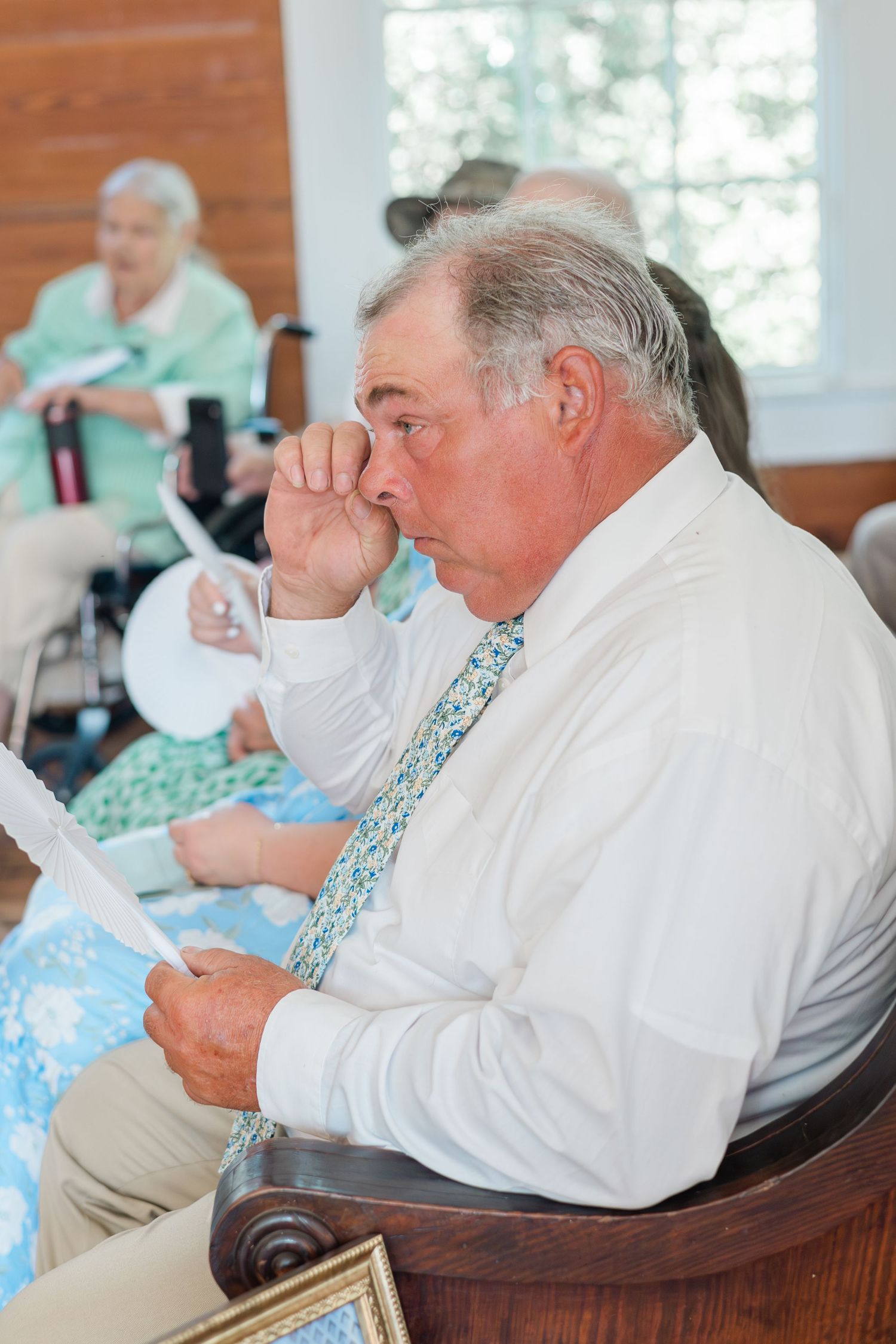 Elderly man in white shirt and floral tie reads paper at senior gathering in bright community room.
