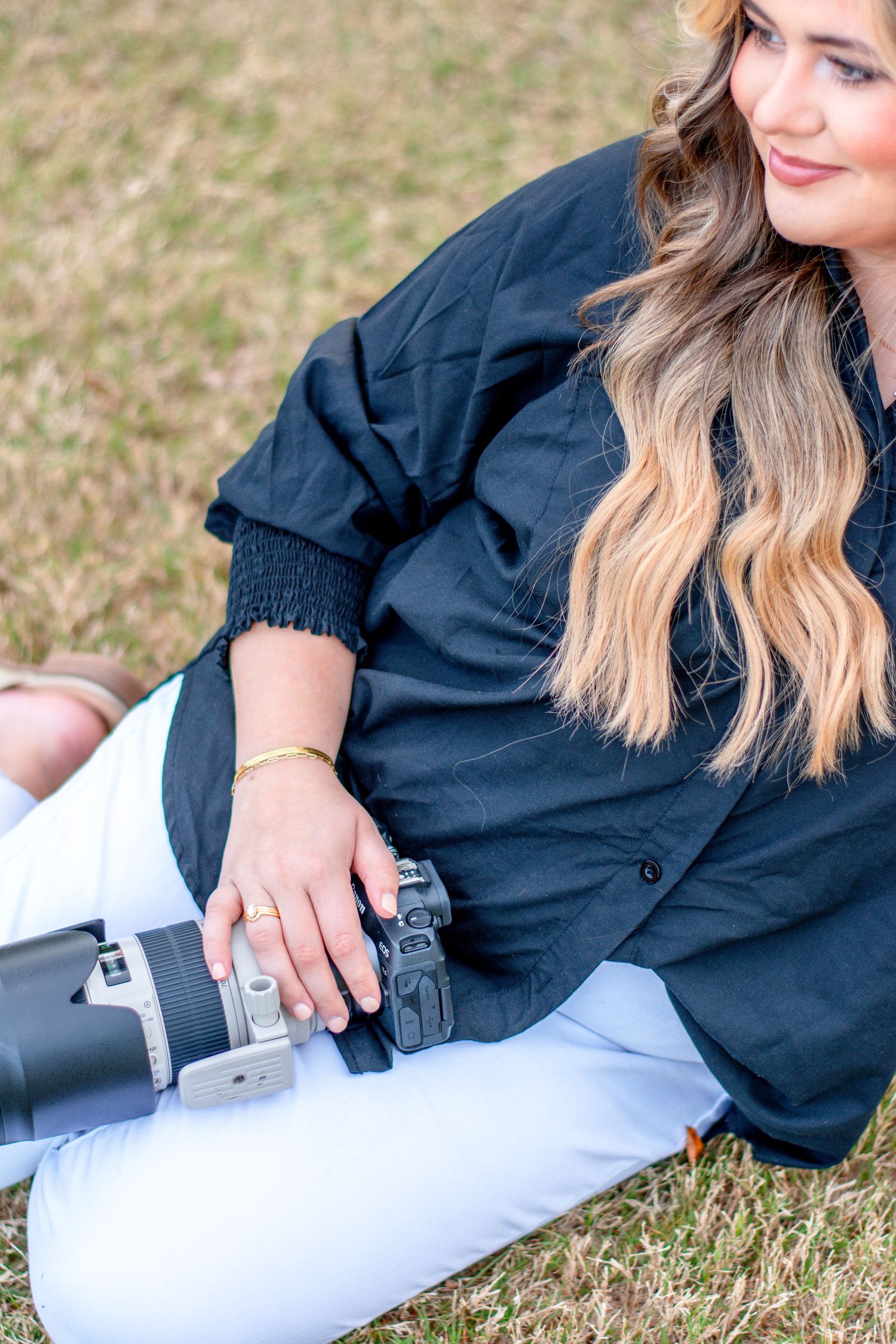 Woman in black shirt and white pants sitting on grass holding a professional camera with telephoto lens.
