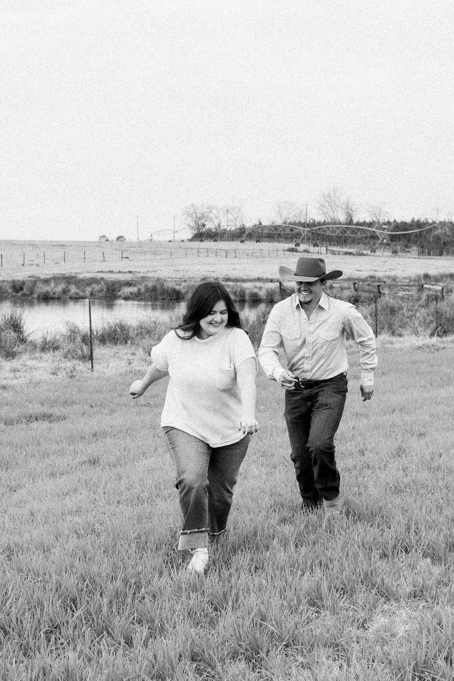 A couple running together through a grassy meadow with a lake in the background.