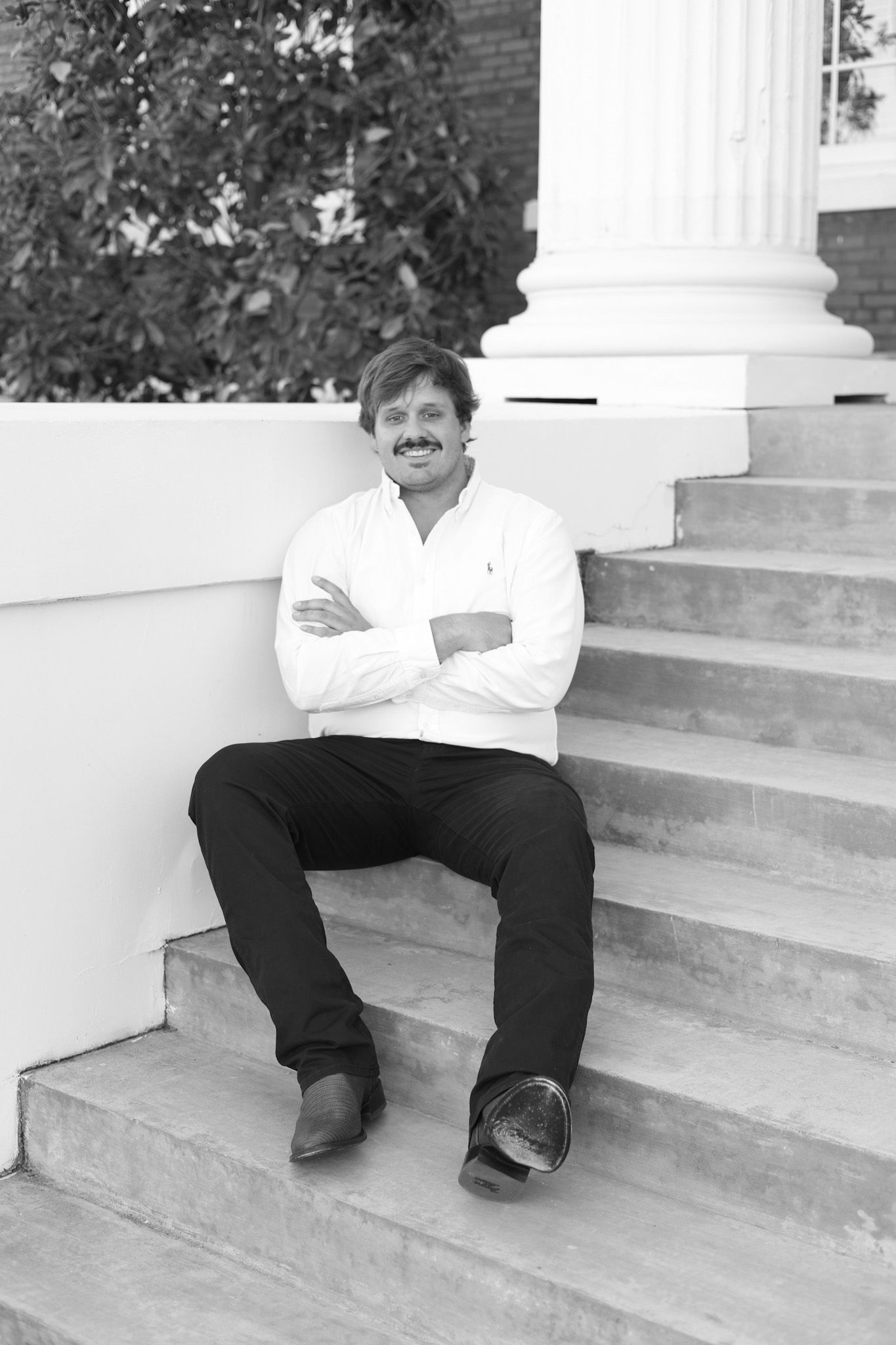 Graduate in cap and gown sitting on steps of building in black and white portrait.