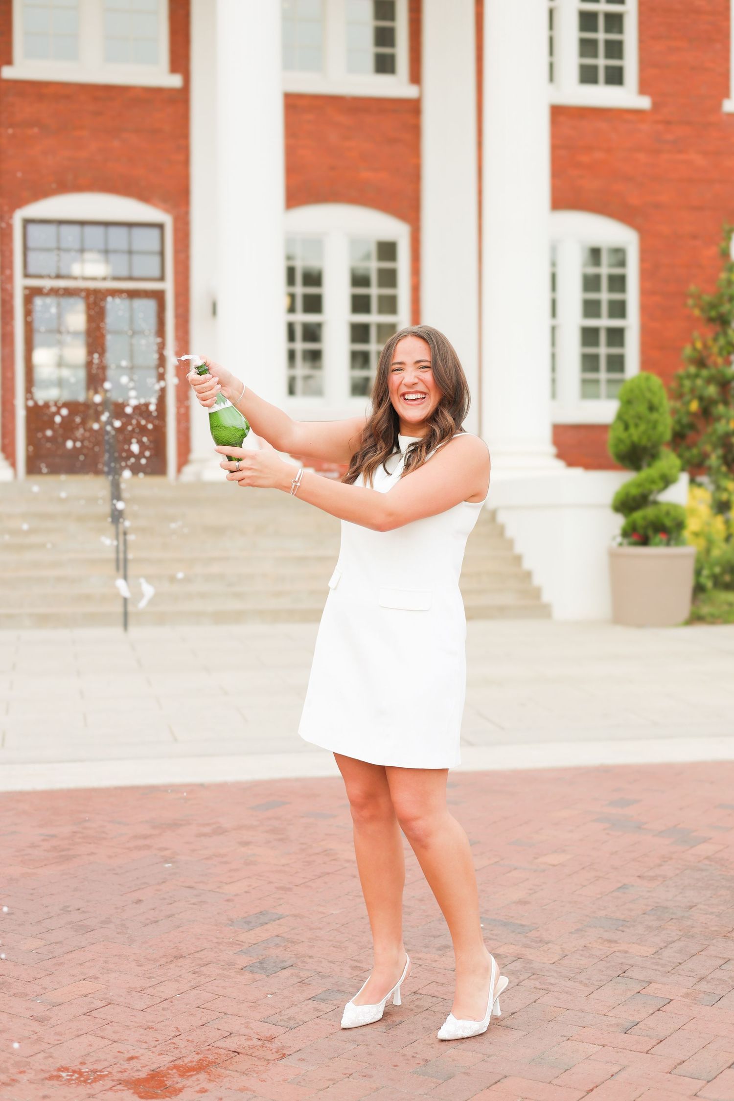 Person in white dress popping champagne bottle in front of red brick building with columns.