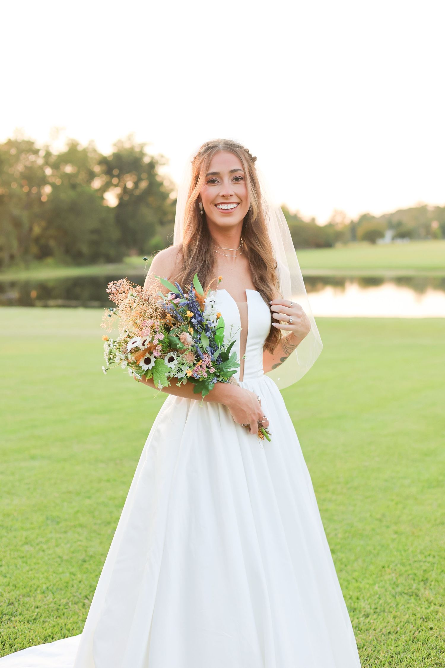 Bride in flowing white dress holds wildflower bouquet while standing in sunlit grassy field during sunset.