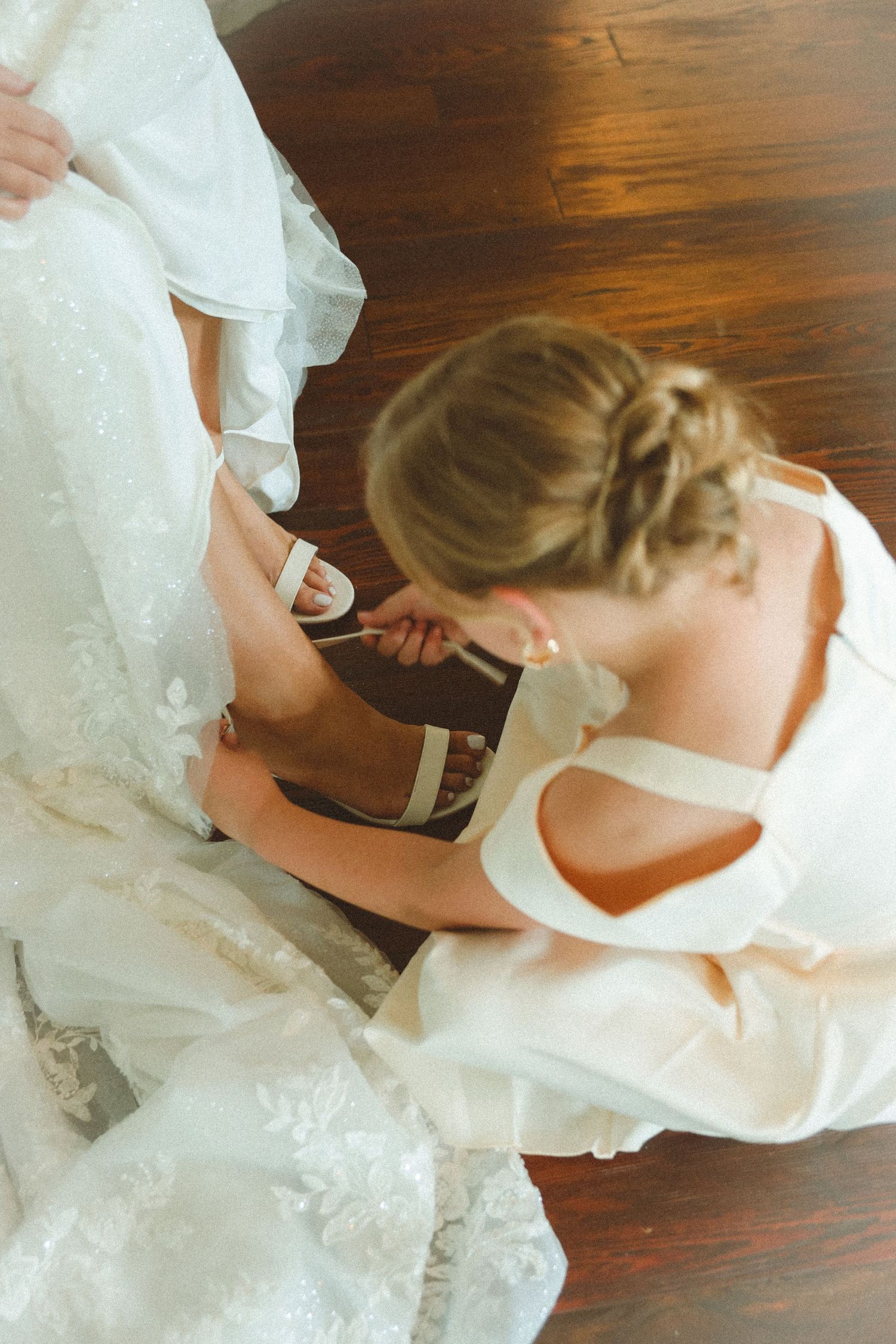 Bridesmaid helping bride put on white heeled wedding shoe while sitting on hardwood floor in elegant gown.