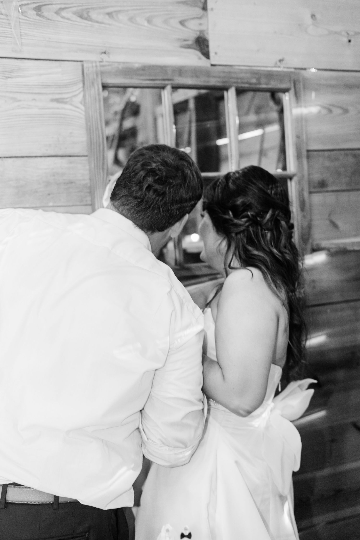 Black and white photo of bride and groom sharing an intimate moment near a rustic wooden wall and window.