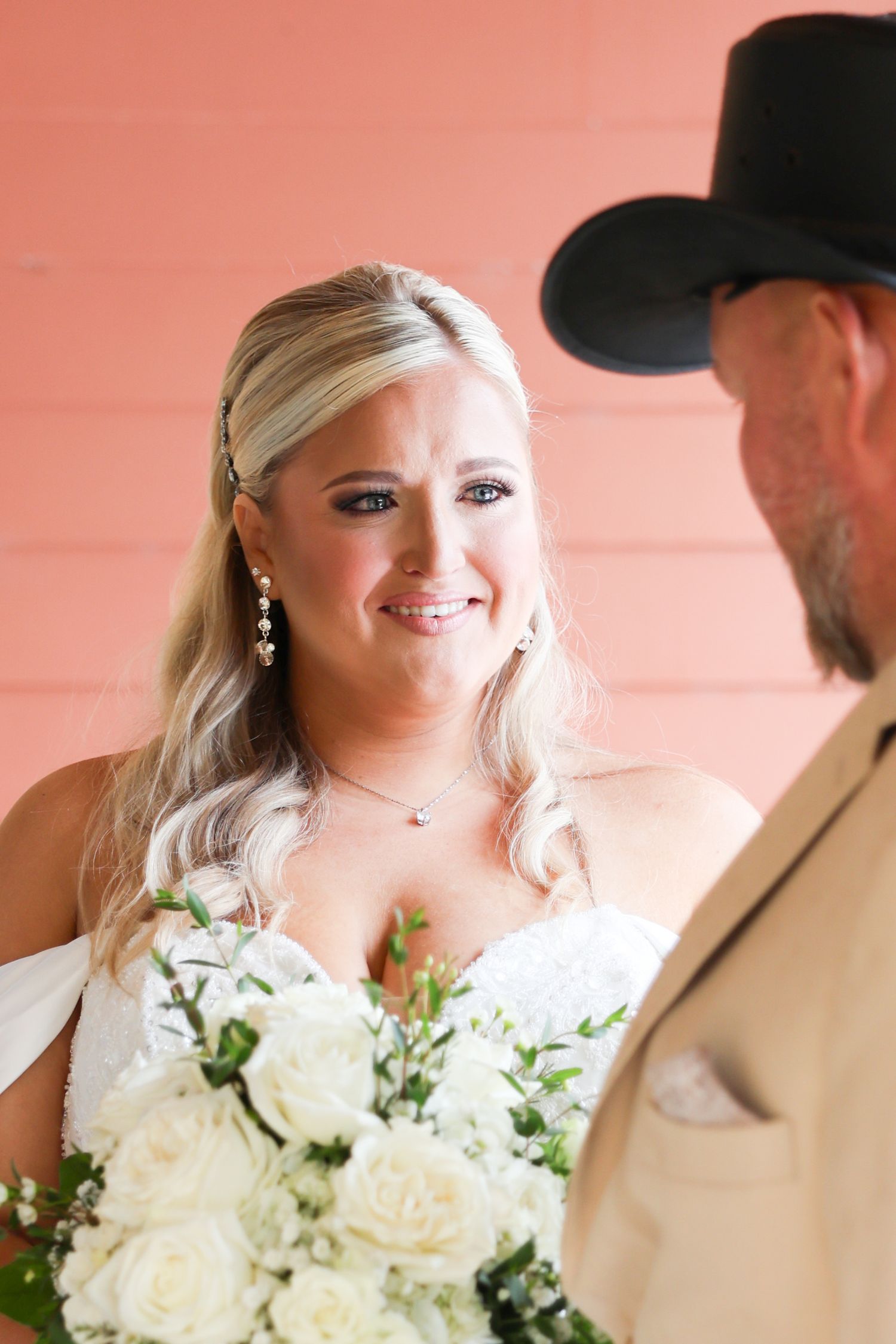 Emotional bride in white gown holding white roses bouquet smiling at groom in cowboy hat during wedding ceremony.
