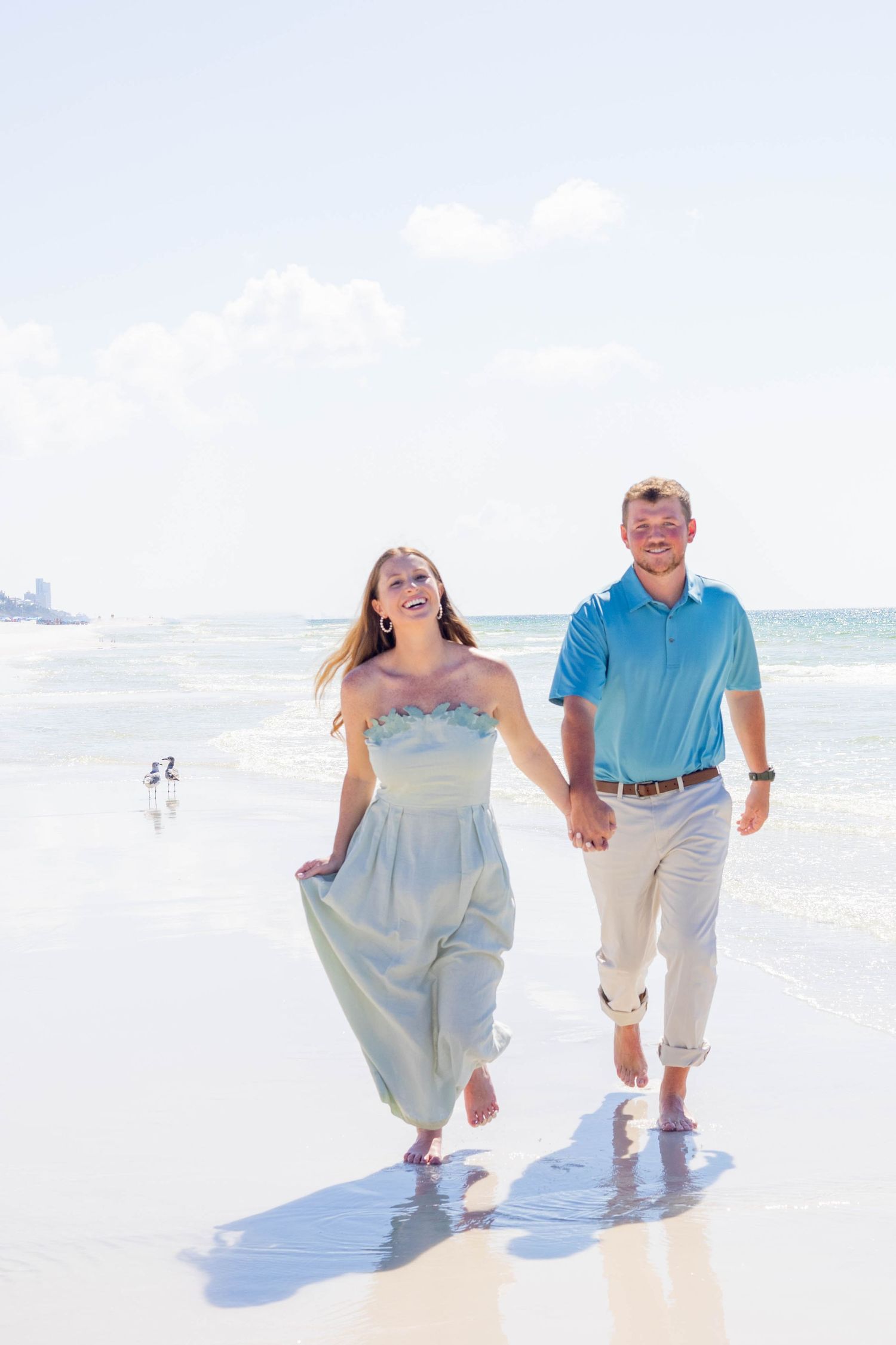 Happy couple walking hand-in-hand barefoot on a sunny beach, woman in light blue dress, man in turquoise polo shirt.