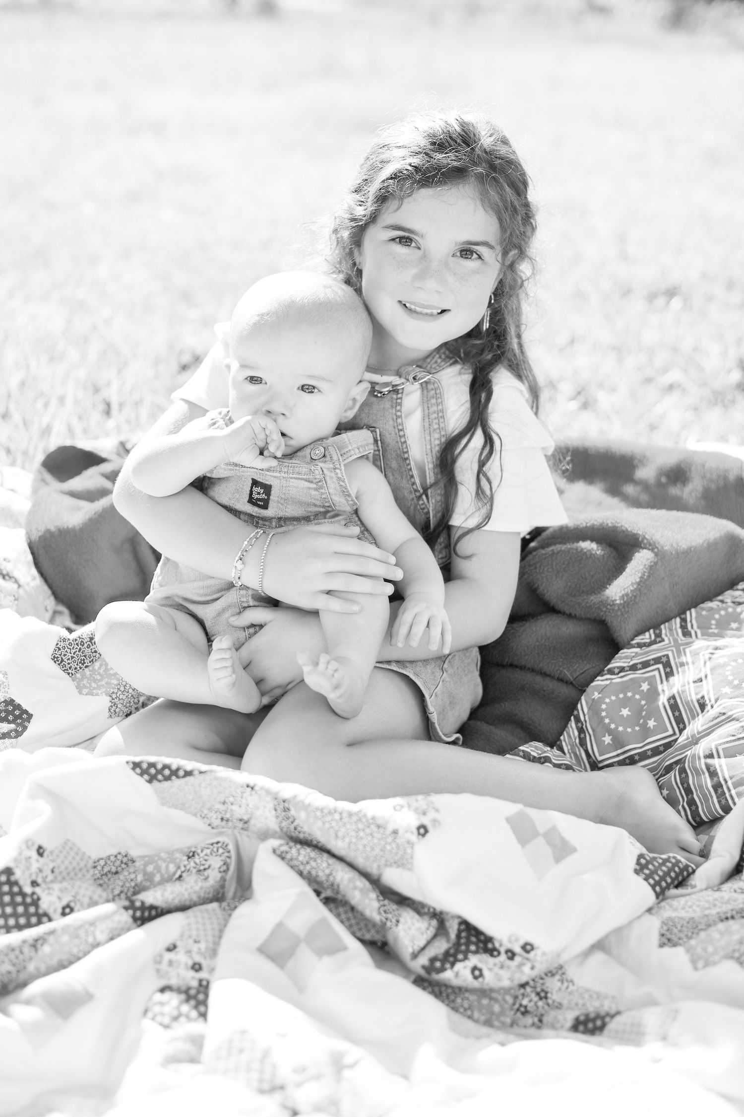Black and white photo of a smiling girl holding a baby on a patchwork quilt outdoors.