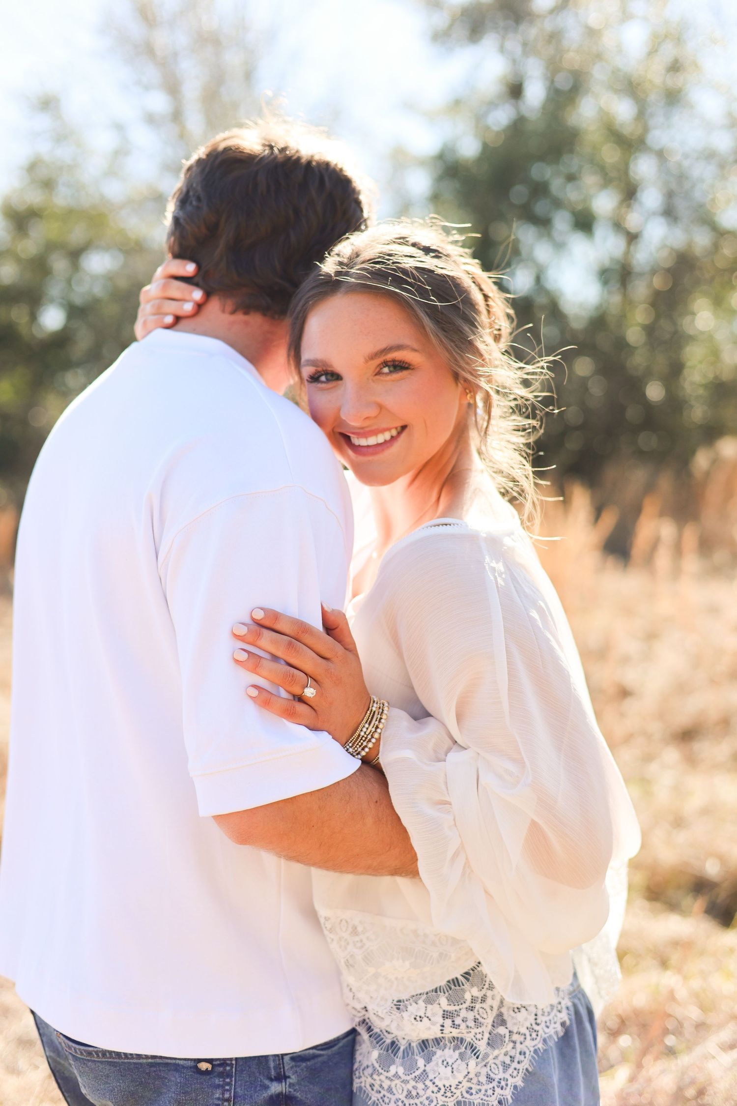A couple in white outfits share intimate embraces in a sun-drenched outdoor setting with golden grass in the background.