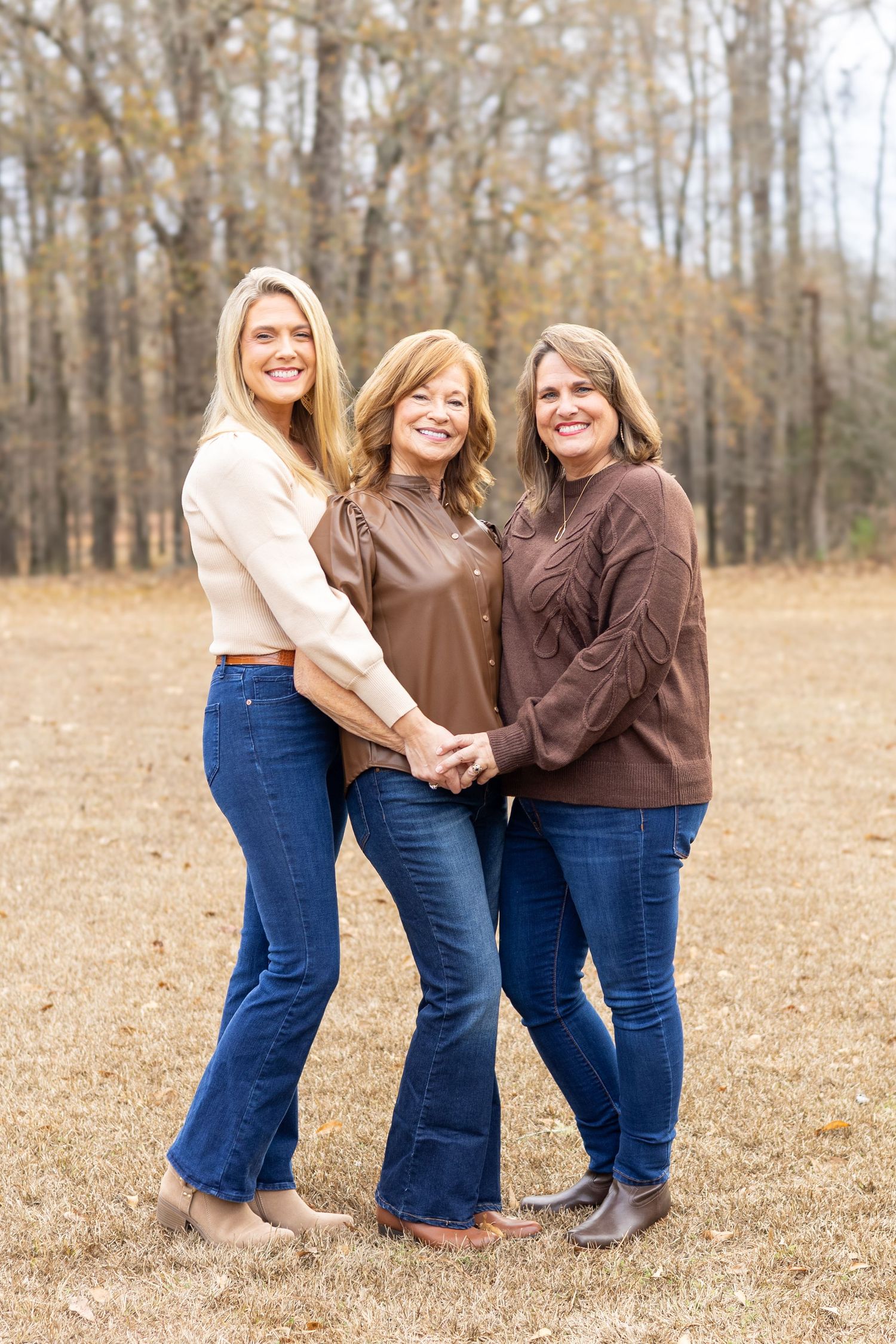 Three people in casual sweaters and jeans pose together outdoors in a fall field with trees in the background.