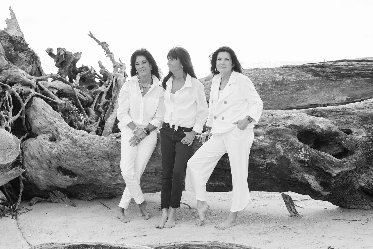 Three people in white outfits sitting on driftwood at the beach in a black and white photo.