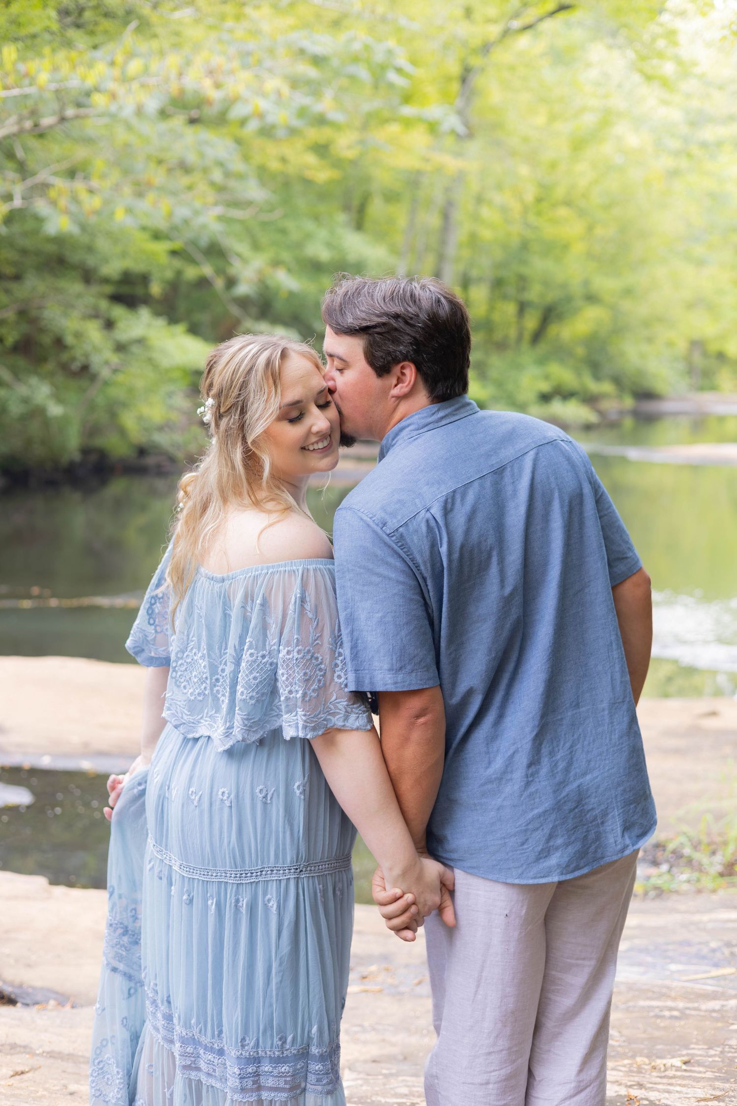 A couple shares a tender kiss outdoors near a creek, woman in blue lace dress, man in blue shirt holding hands.