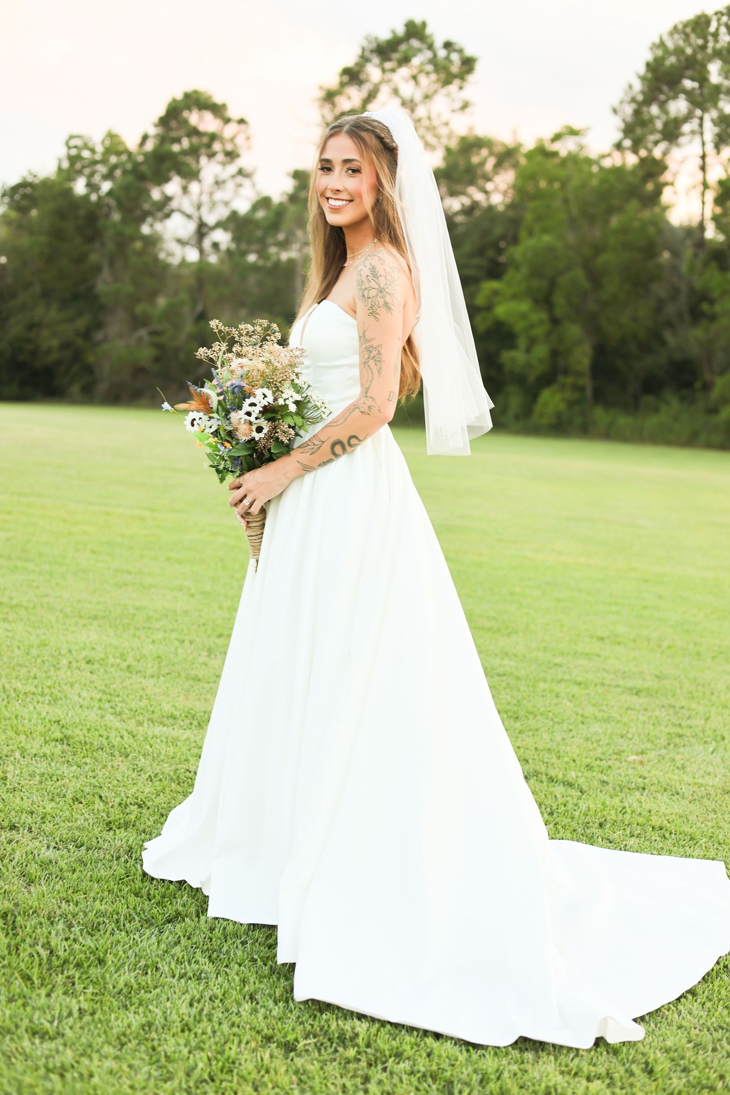 A bride in a ballgown style dress stands in a green field holding a rustic bouquet.