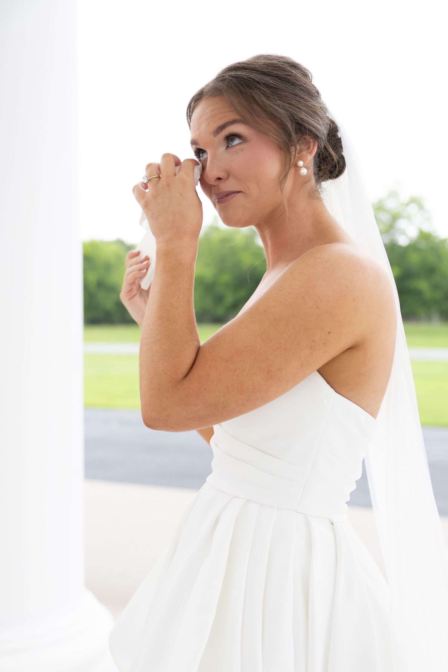 Emotional bride in white strapless gown and veil wiping tears, standing between white columns outdoors.