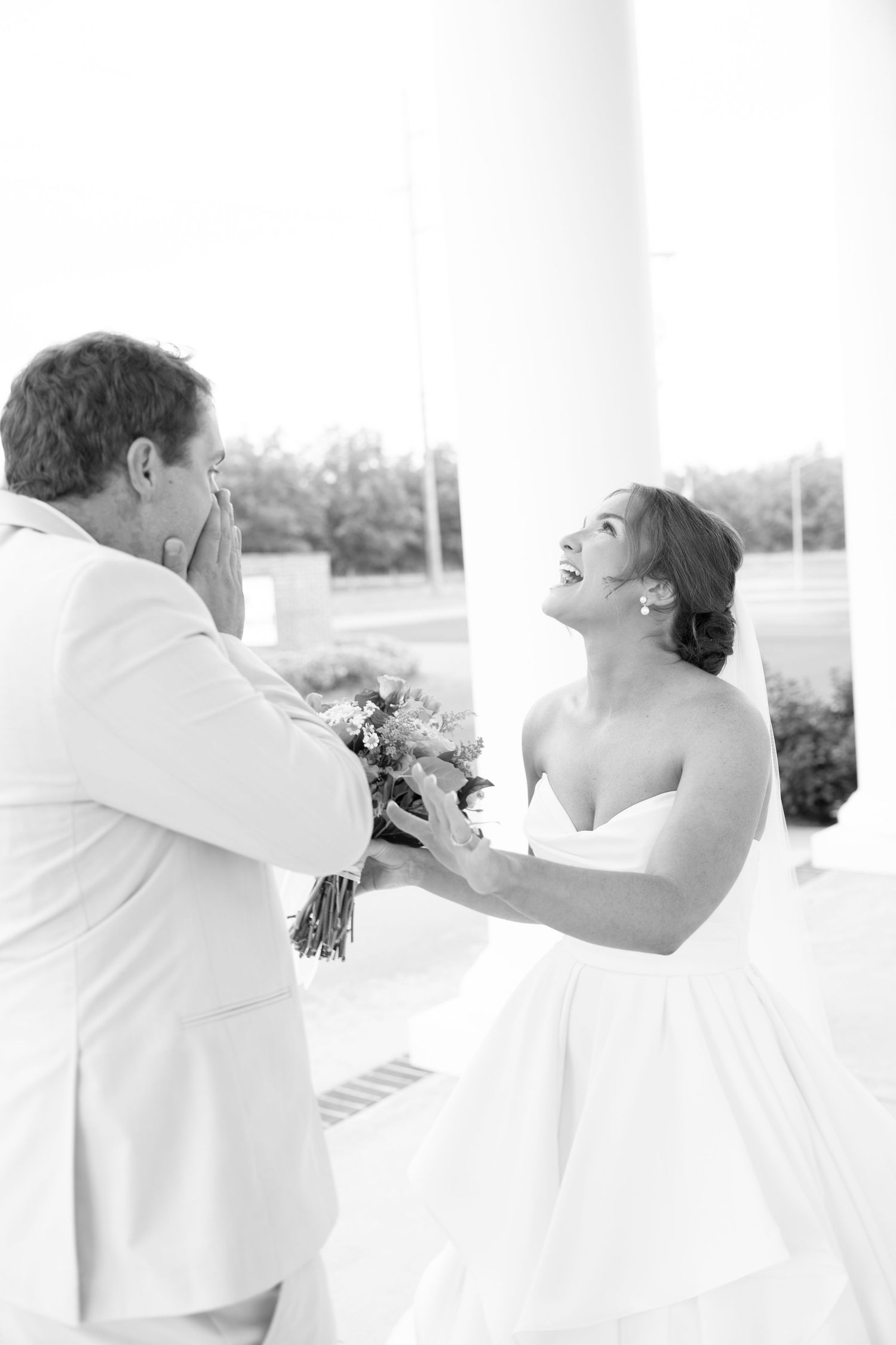 Black and white photo of a bride and groom sharing an emotional first look moment, bride laughing joyfully holding bouquet.
