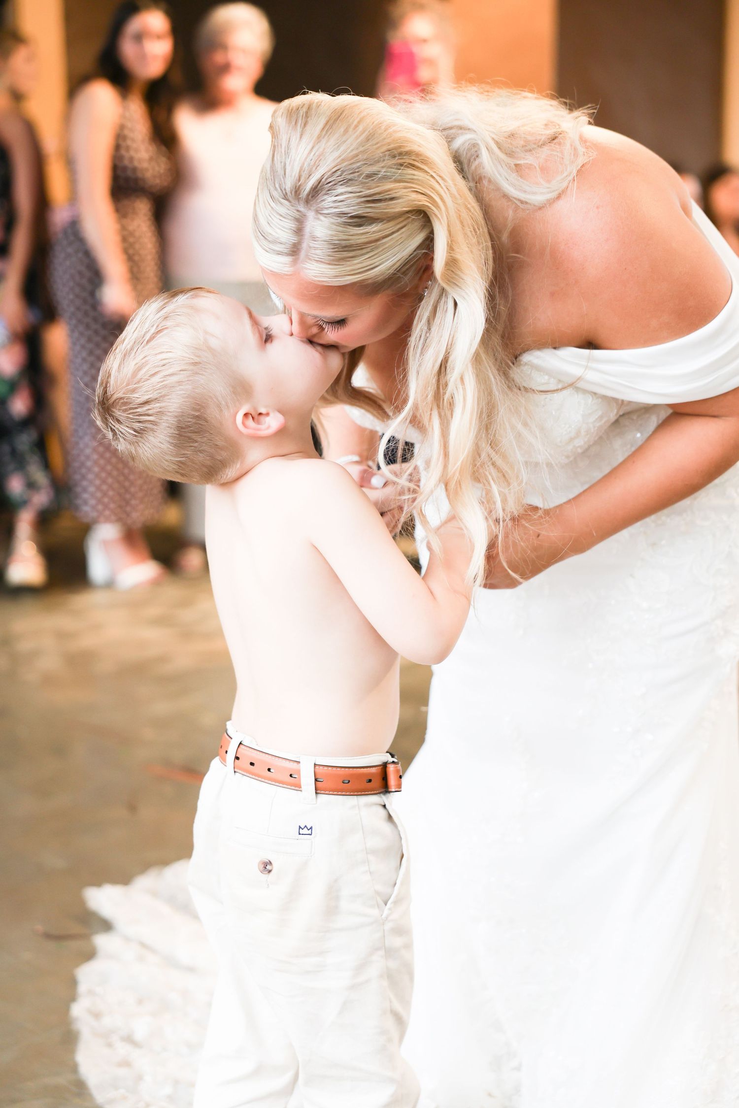 Bride in off-shoulder white wedding gown sharing a sweet kiss with young shirtless ring bearer at reception.