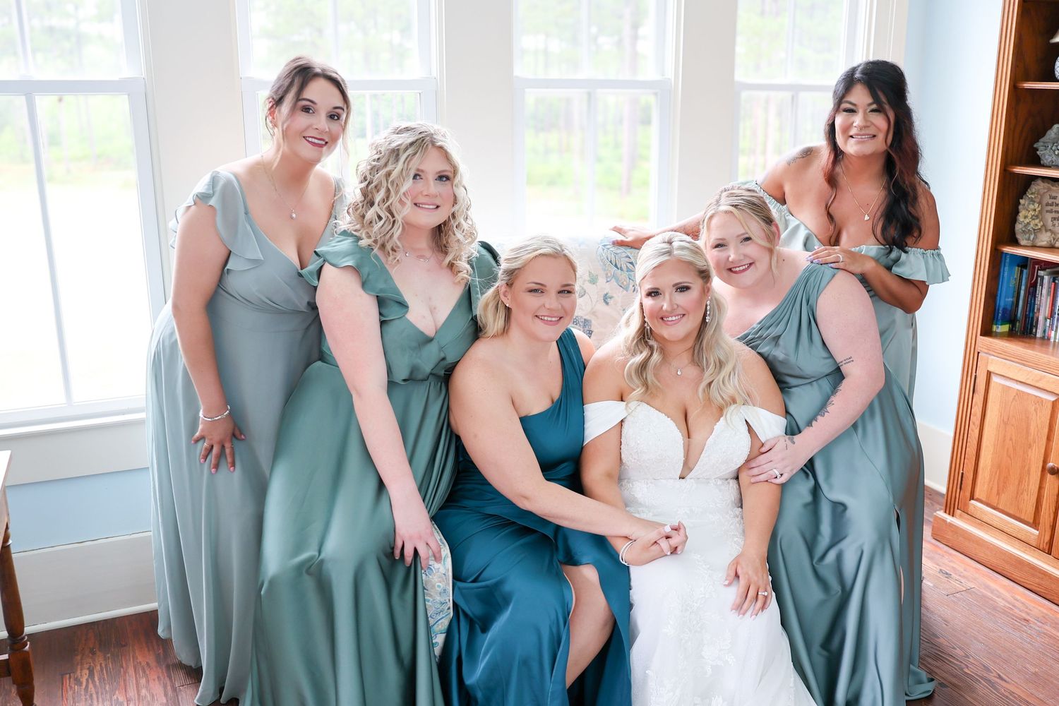 Bride in white gown surrounded by five bridesmaids in sage green dresses posing together indoors before wedding.