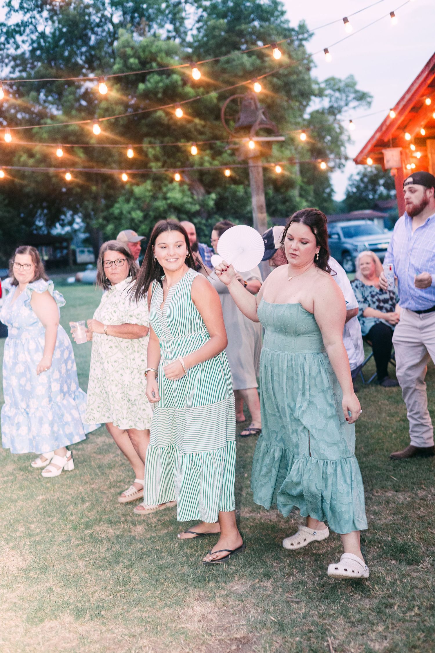 Wedding guests in green and floral dresses dancing under string lights at outdoor reception.
