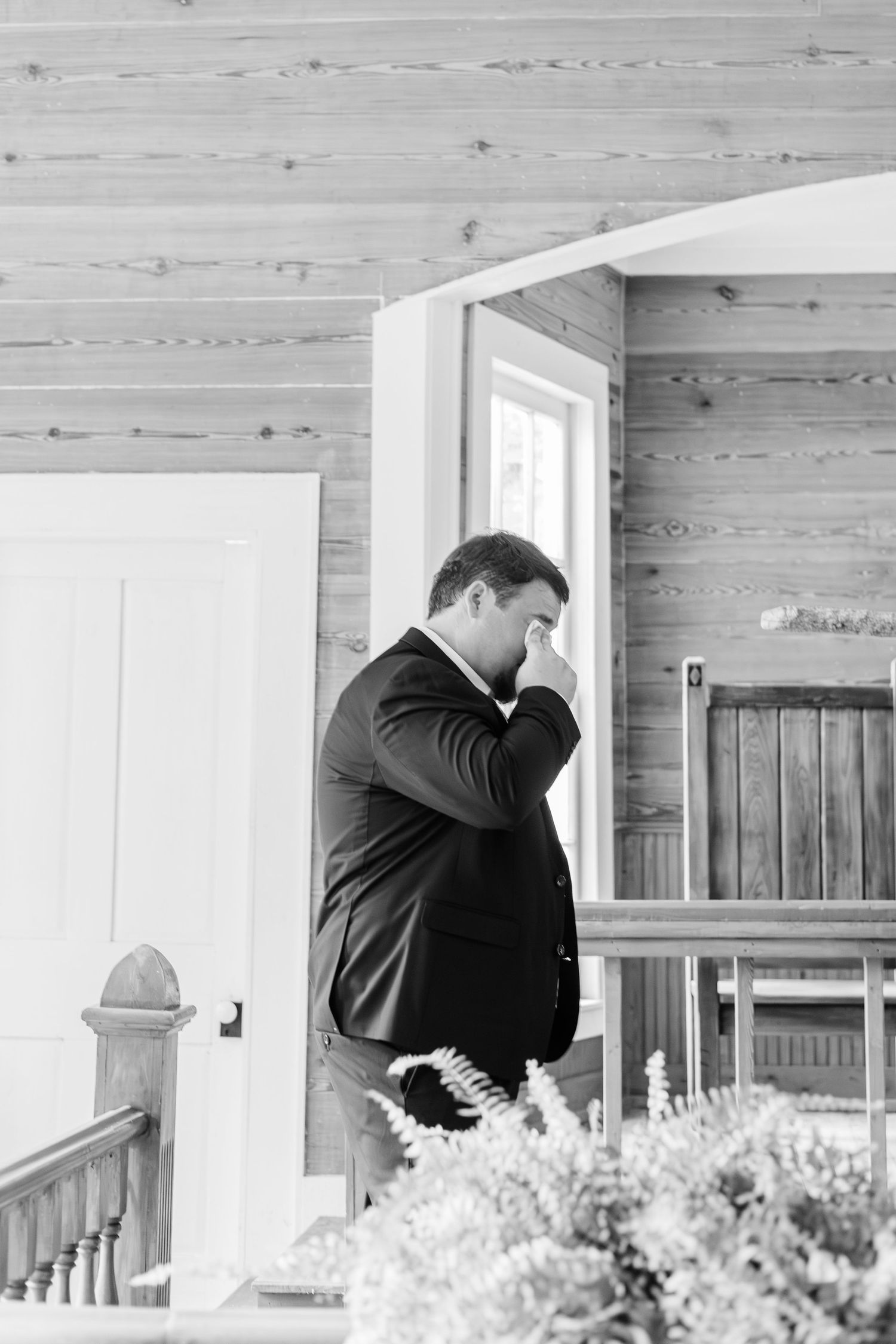 Black and white photo of a groom in a suit standing pensively on a wooden porch before his wedding.