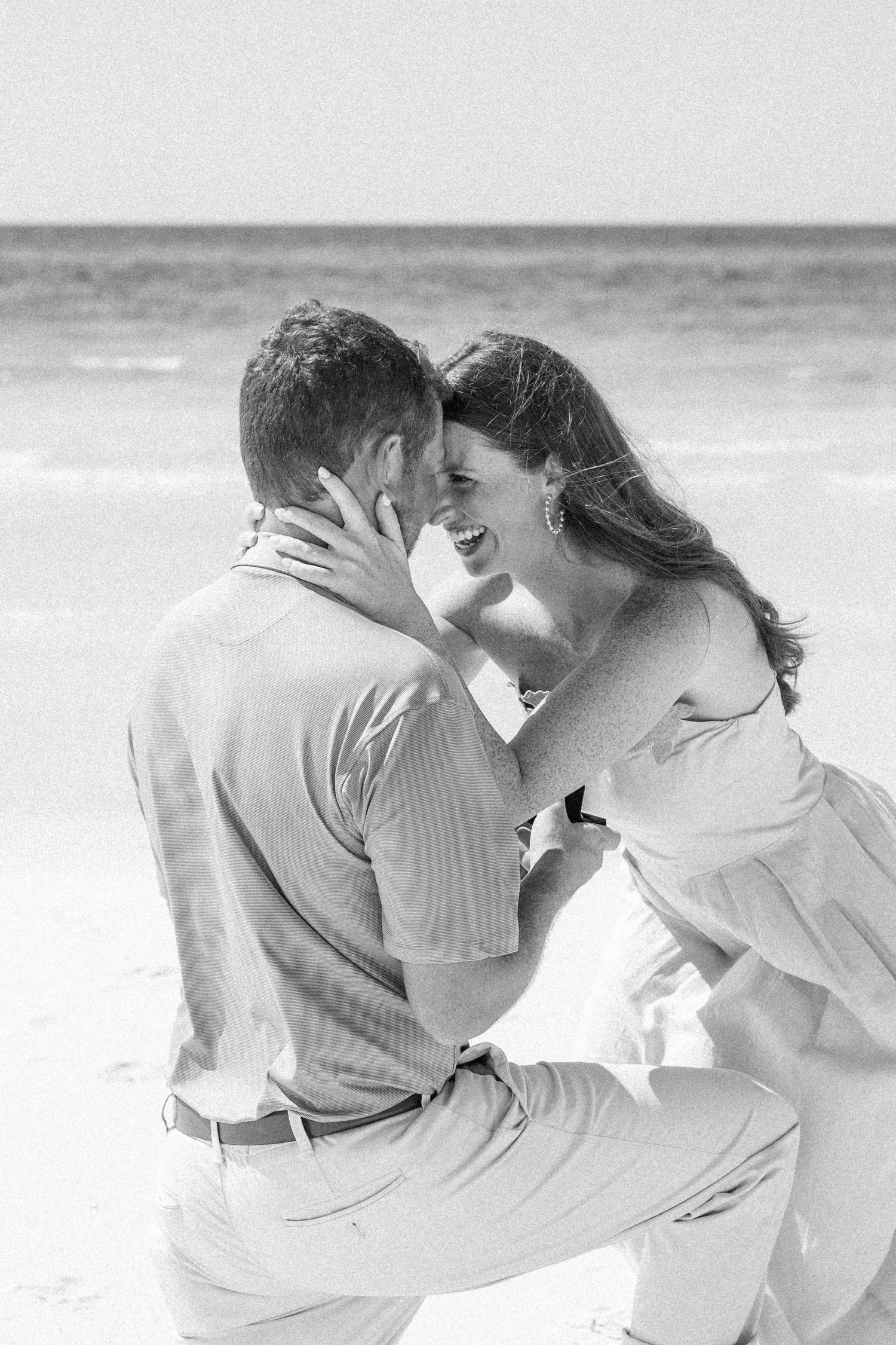 Joyful couple sharing an intimate moment as man kneels before woman during beach marriage proposal.