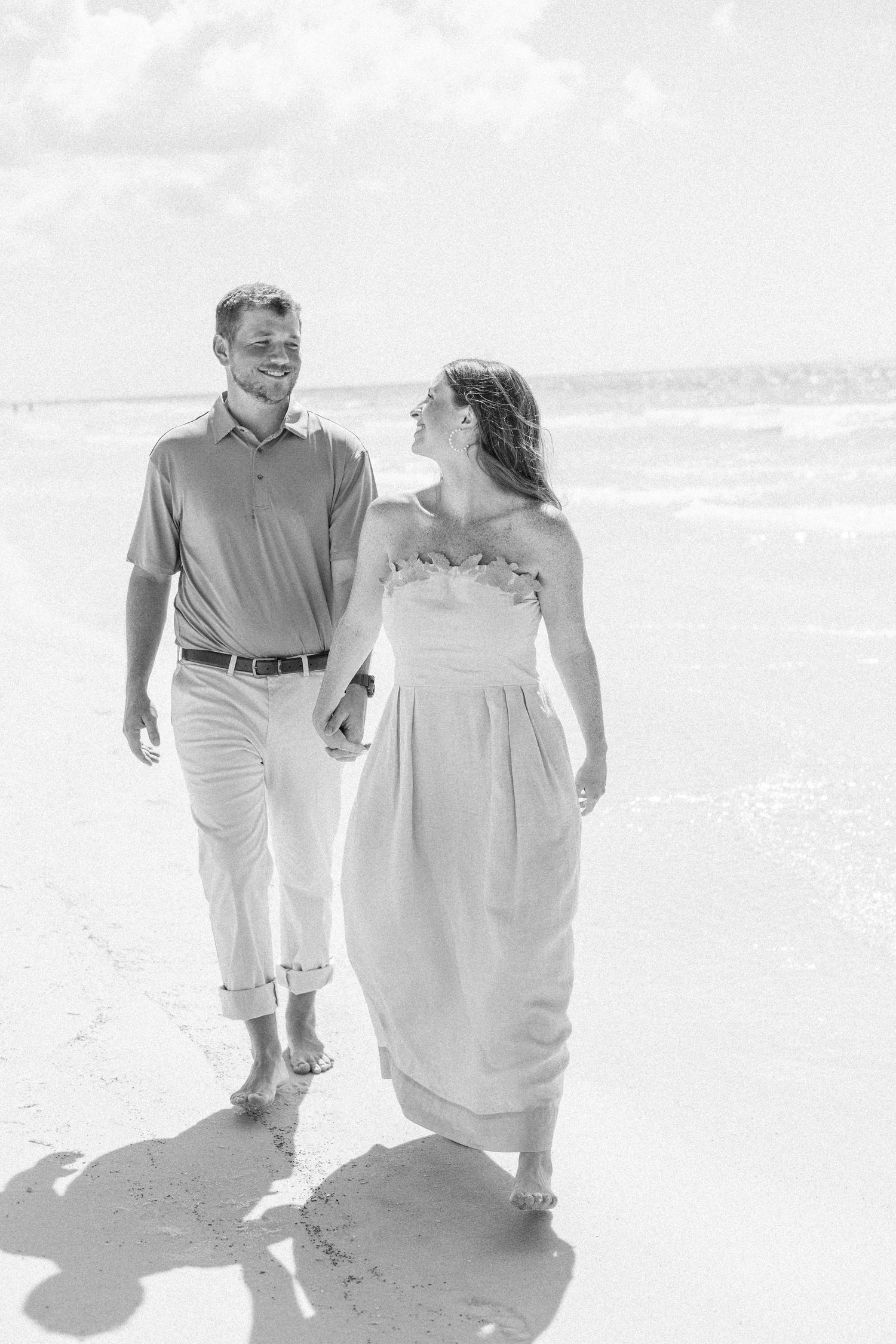 Smiling couple holding hands and walking along a sunny beach shore during engagement photo session.