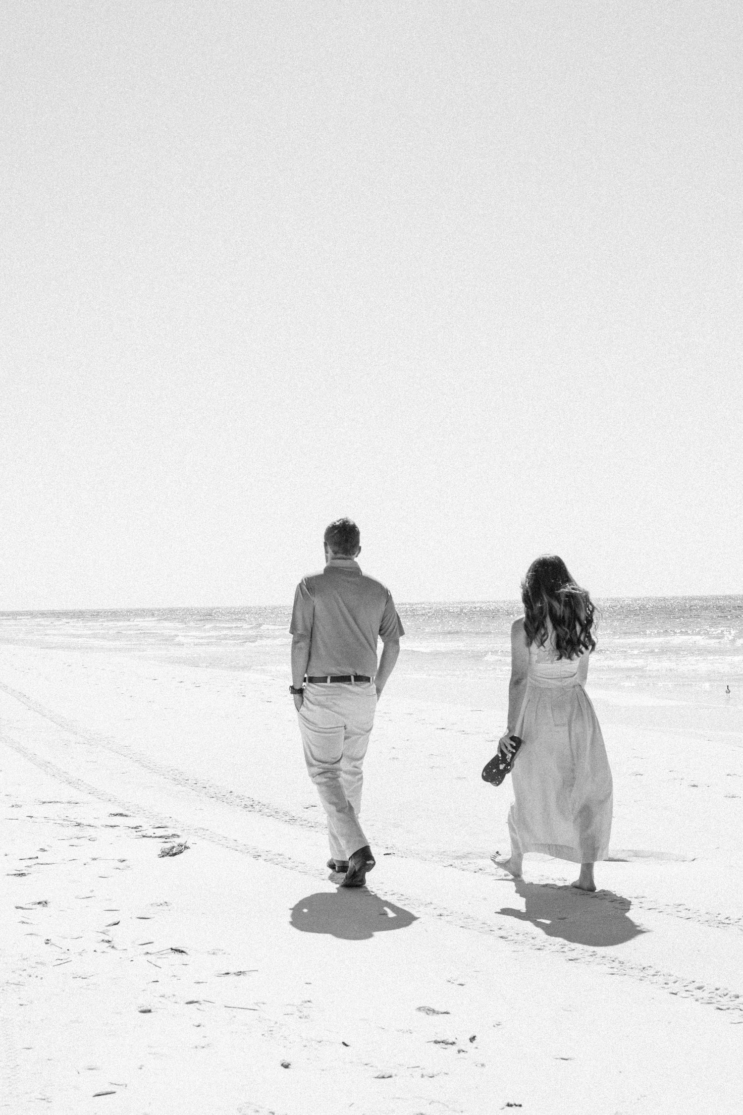 Black and white photo of a couple walking hand in hand on a sandy beach toward the ocean.