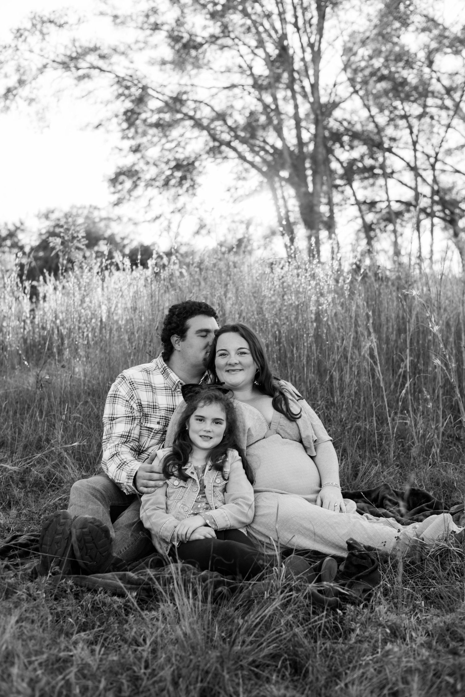 Black and white photo of a pregnant woman, man, and young girl sitting together in a field of tall grass.