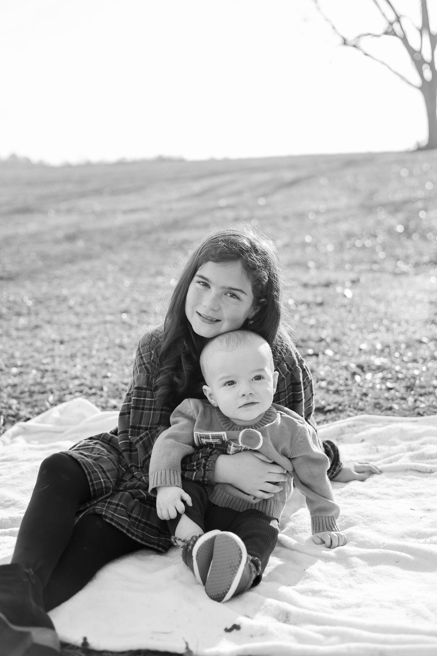 Black and white photo of a young girl holding a baby on a blanket outdoors near a bare tree.